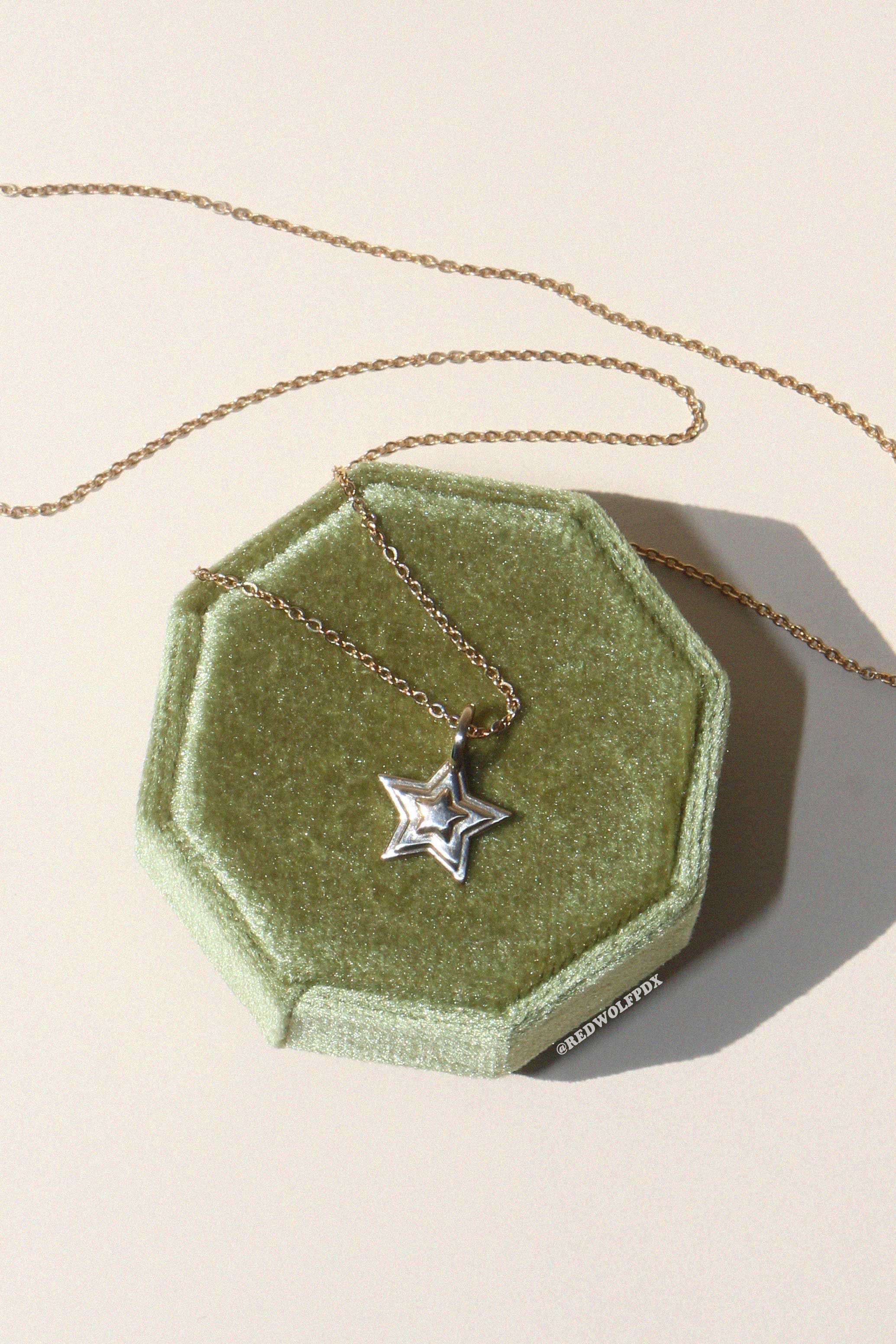 A brass starburst 3-d layered charm hanging from a brass chain necklace displayed on a green octagonal box on a beige background.