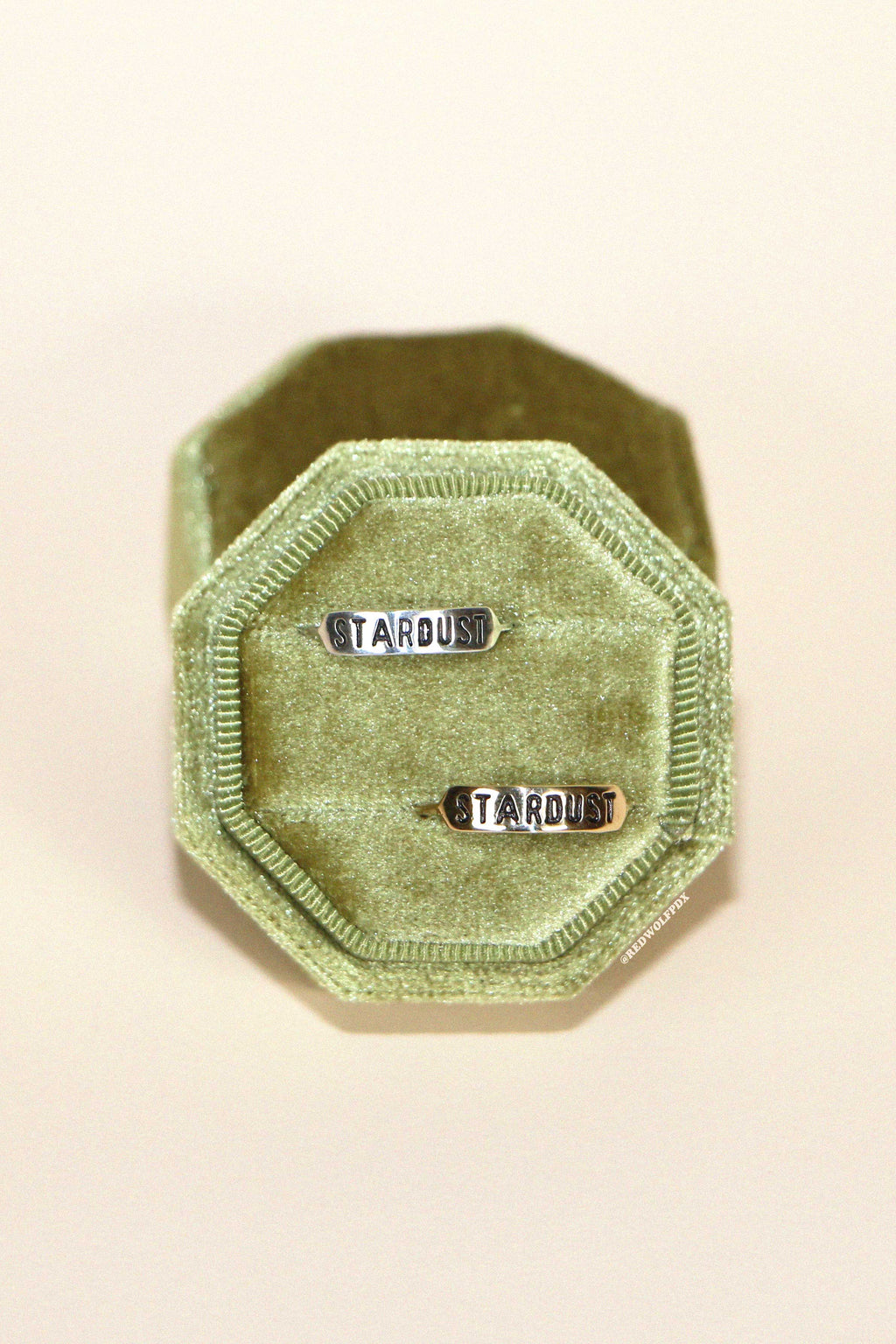 A green velvet ring box  holding a sterling silver ring and a brass ring both with the text 'STARDUST' engraved on them against a beige backdrop