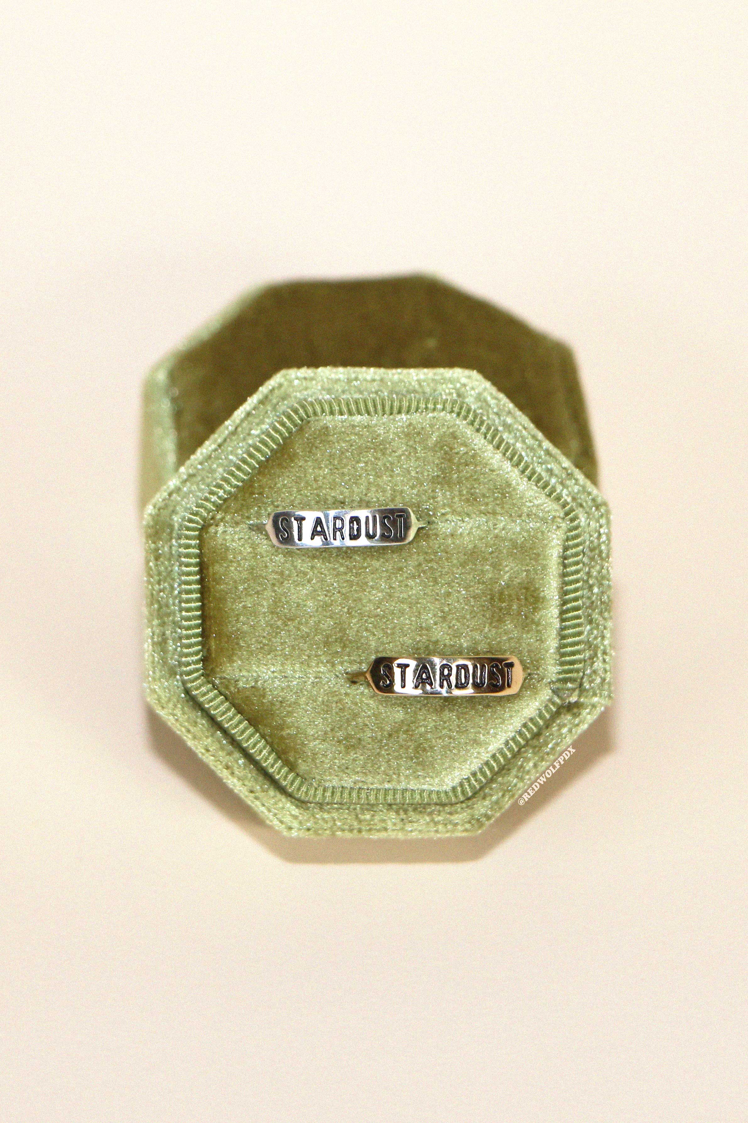 A green velvet ring box  holding a sterling silver ring and a brass ring both with the text 'STARDUST' engraved on them against a beige backdrop