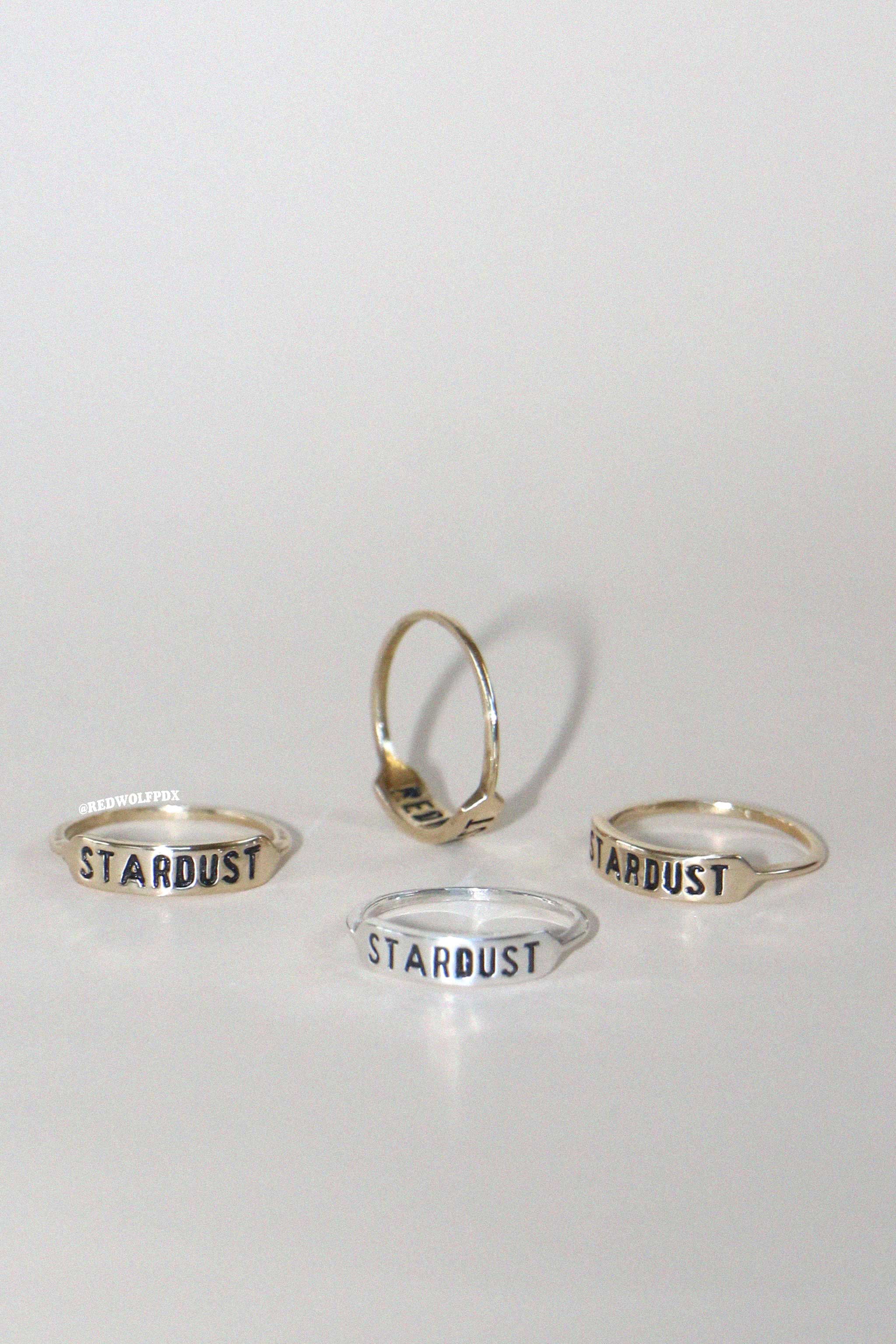 a sterling silver ring and three brass rings all with the text 'STARDUST' engraved on them scattered across a beige backdrop