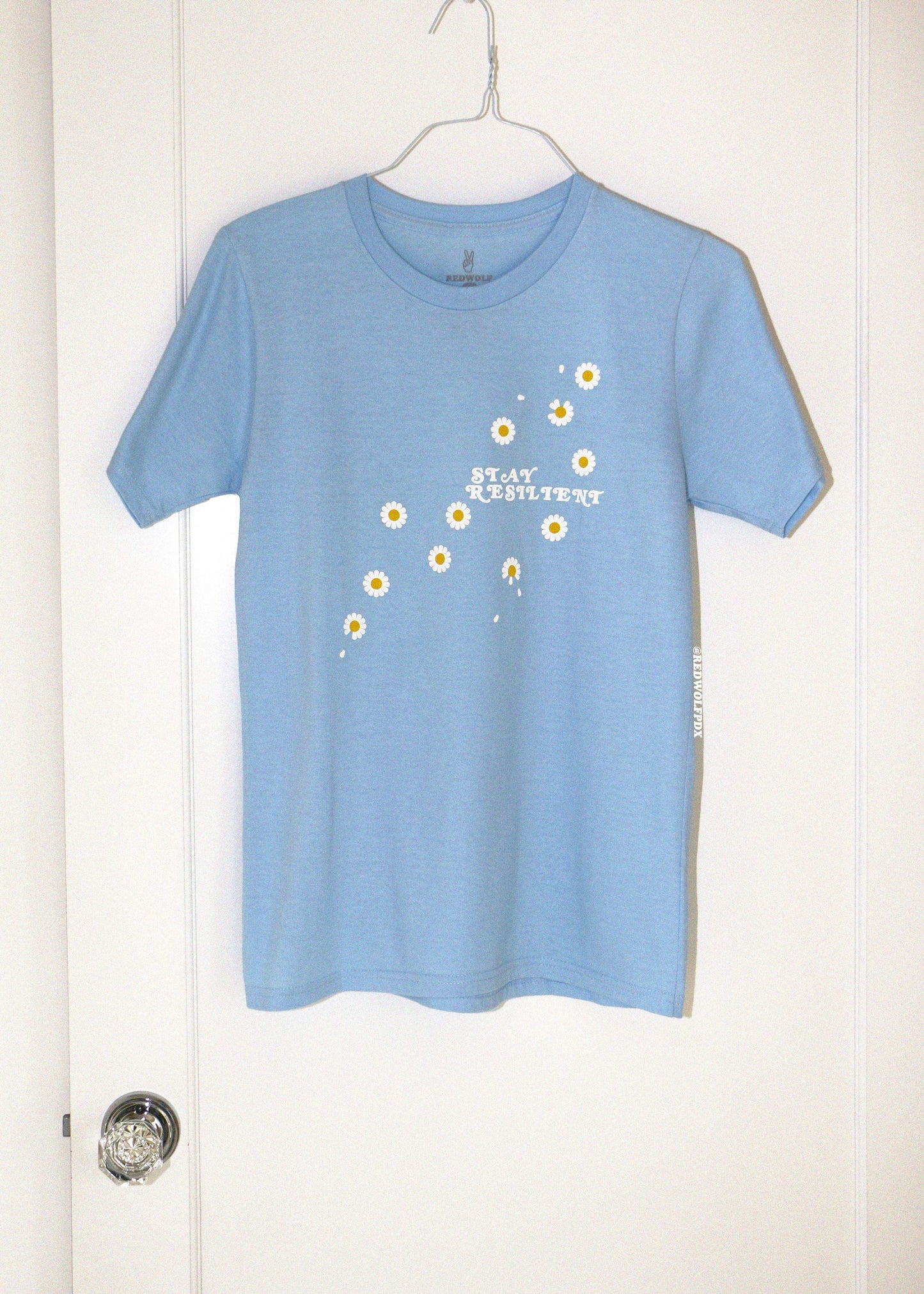 Blue t-shirt with floral design and text on a hanger against a white door.