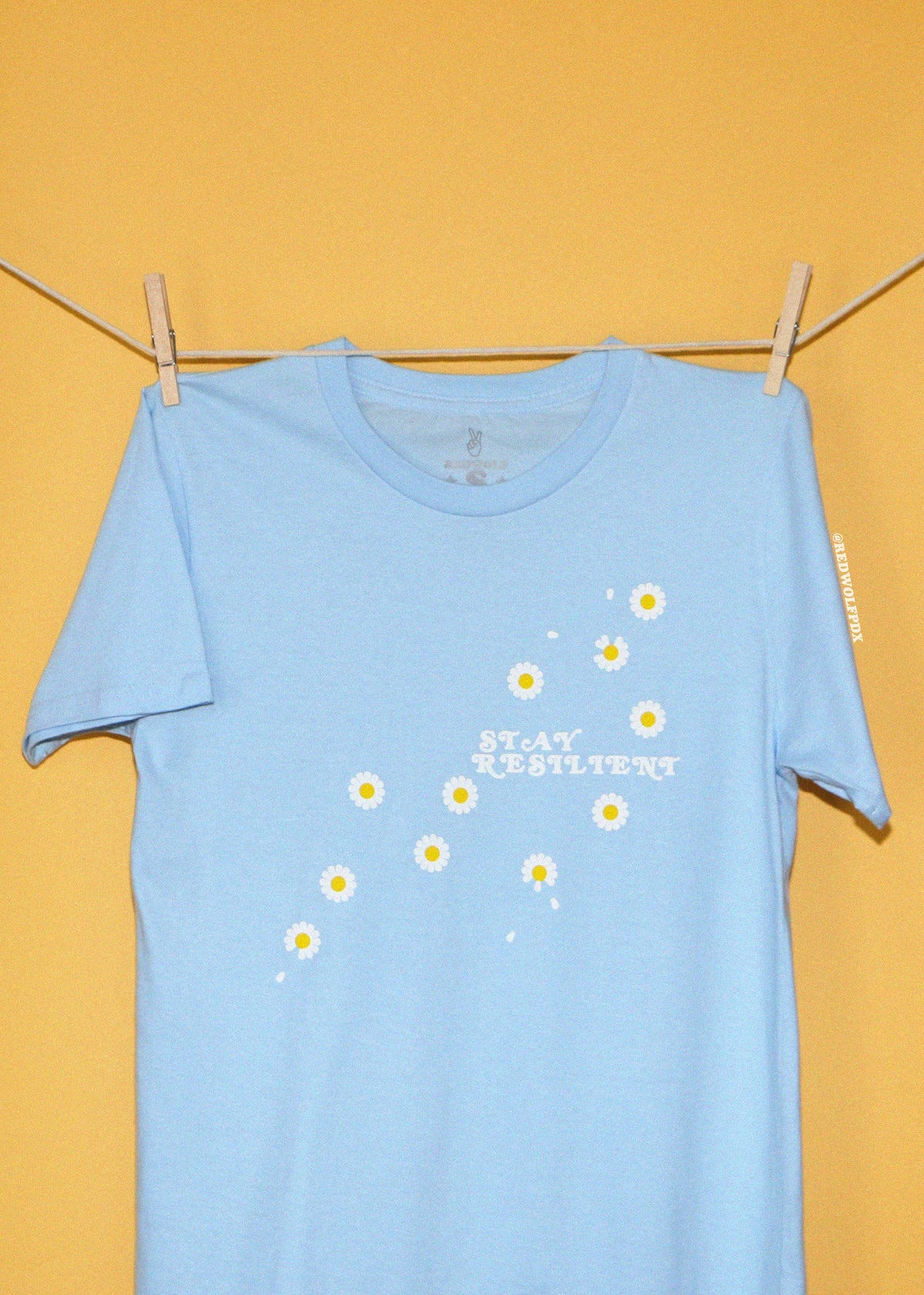 A light blue short-sleeve t-shirt hanging from a wooden clothesline with two wooden clothespins against a solid yellow background. The shirt features a graphic of small white daisies with yellow centers and the text 'STAY RESILIENT' printed in white.