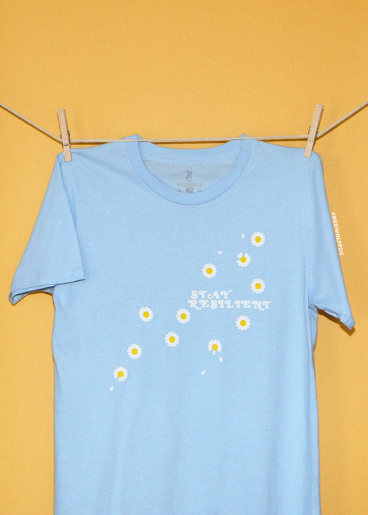 A light blue short-sleeve t-shirt hanging from a wooden clothesline with two wooden clothespins against a solid yellow background. The shirt features a graphic of small white daisies with yellow centers and the text 'STAY RESILIENT' printed in white.