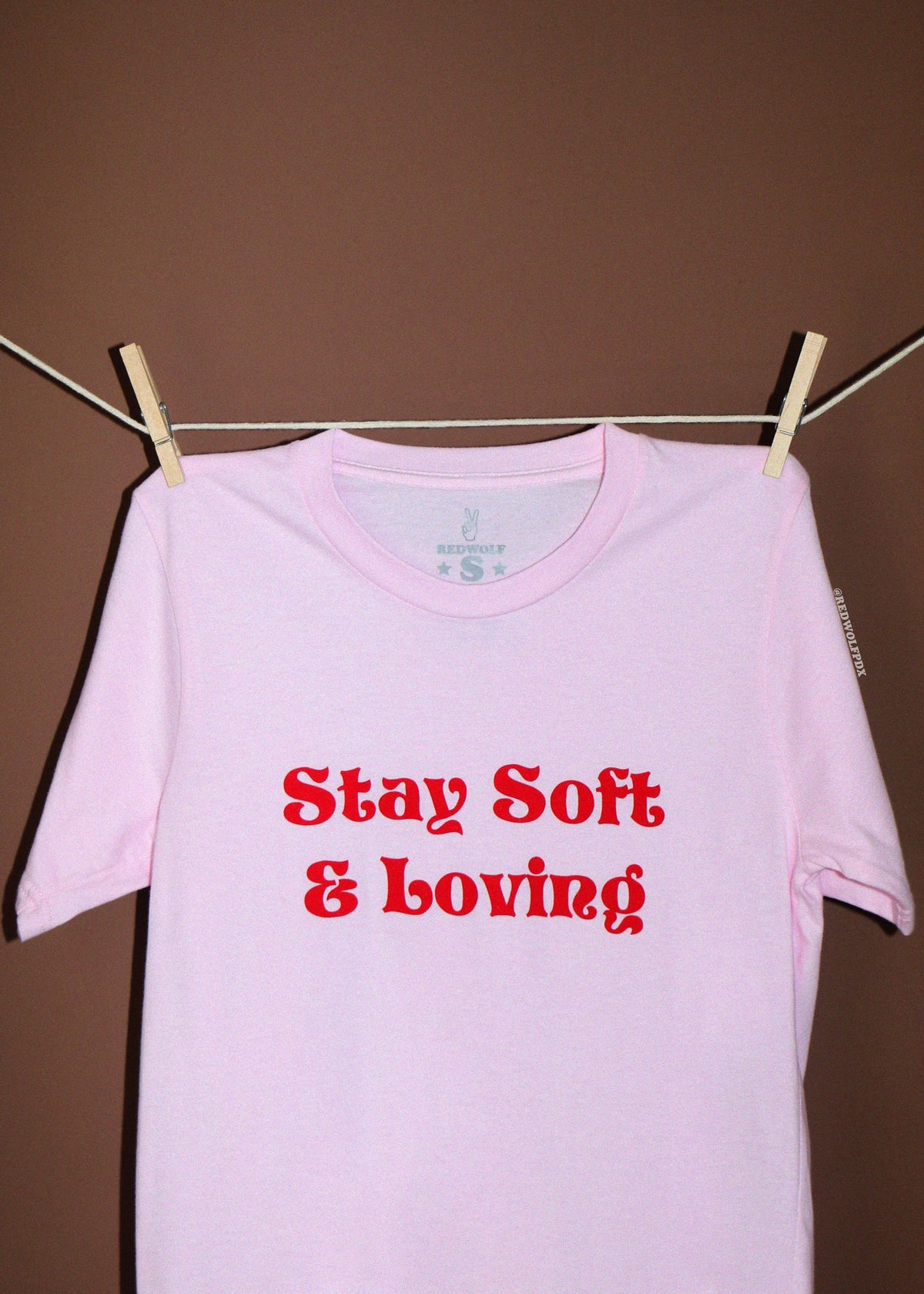 A light pink short-sleeve t-shirt with text 'Stay Soft & Loving' printed in red across the chest, hanging from a clothesline with wooden clothespins against a solid brown background.