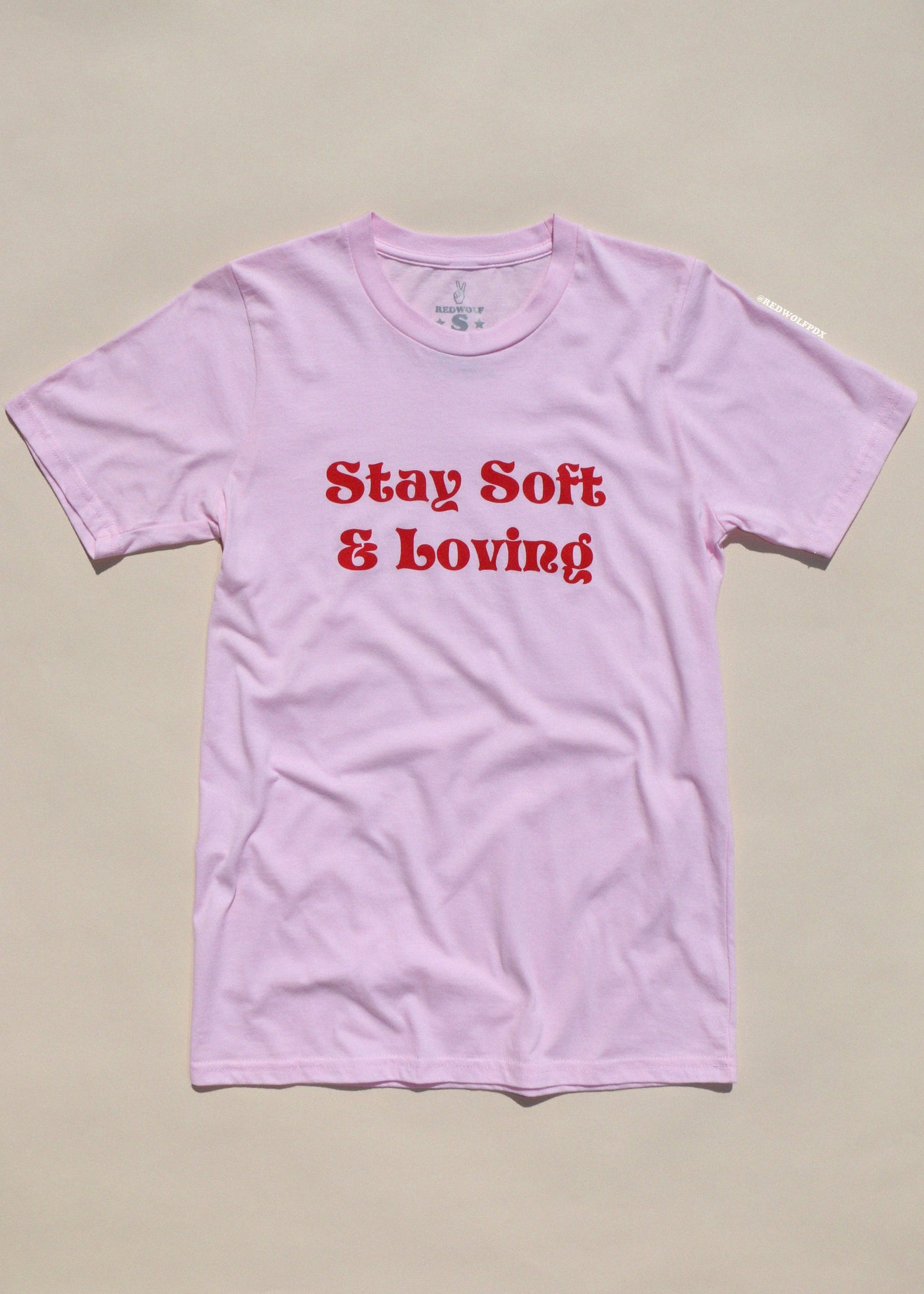 Pink t-shirt with red text 'Stay Soft & Loving' on a beige background