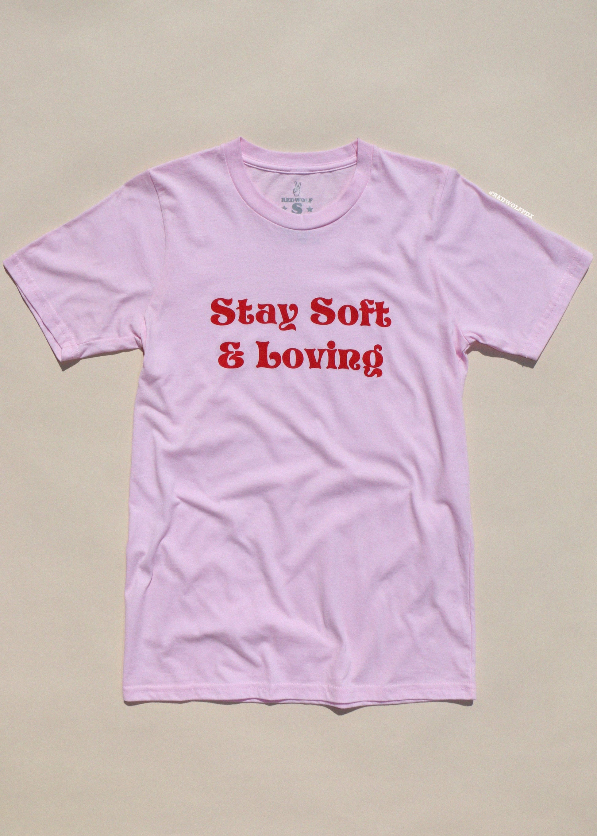 Pink t-shirt with red text 'Stay Soft & Loving' on a beige background