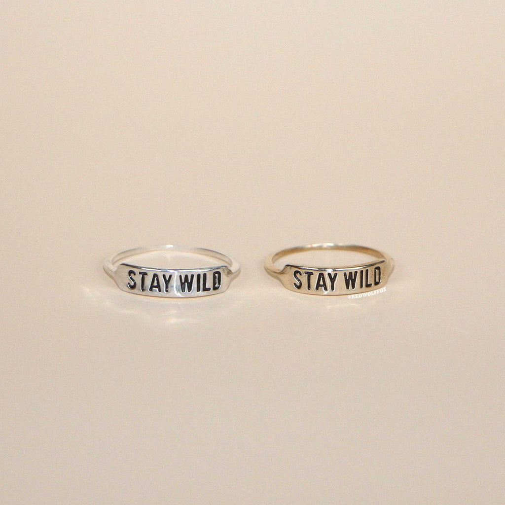 Two rings, one engraved with 'STAY WILD' on a silver finish and the other on a brass finish, placed side by side on a neutral background.