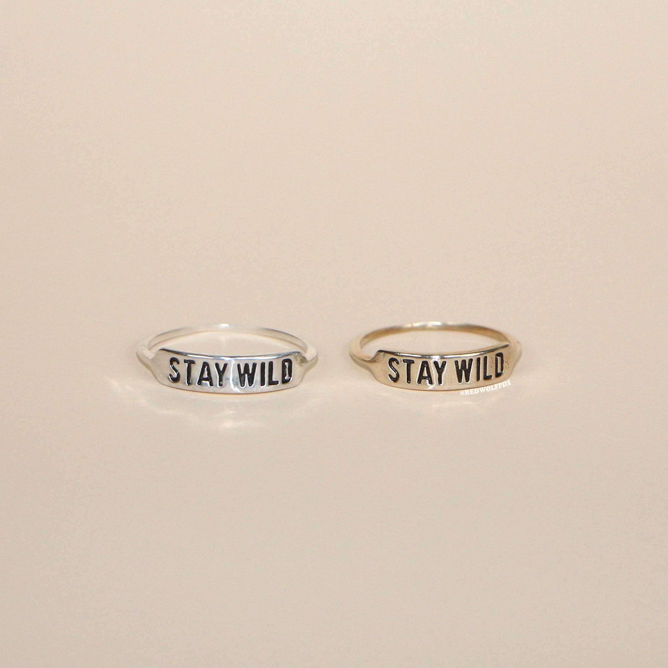 Two rings, one engraved with 'STAY WILD' on a silver finish and the other on a brass finish, placed side by side on a neutral background.