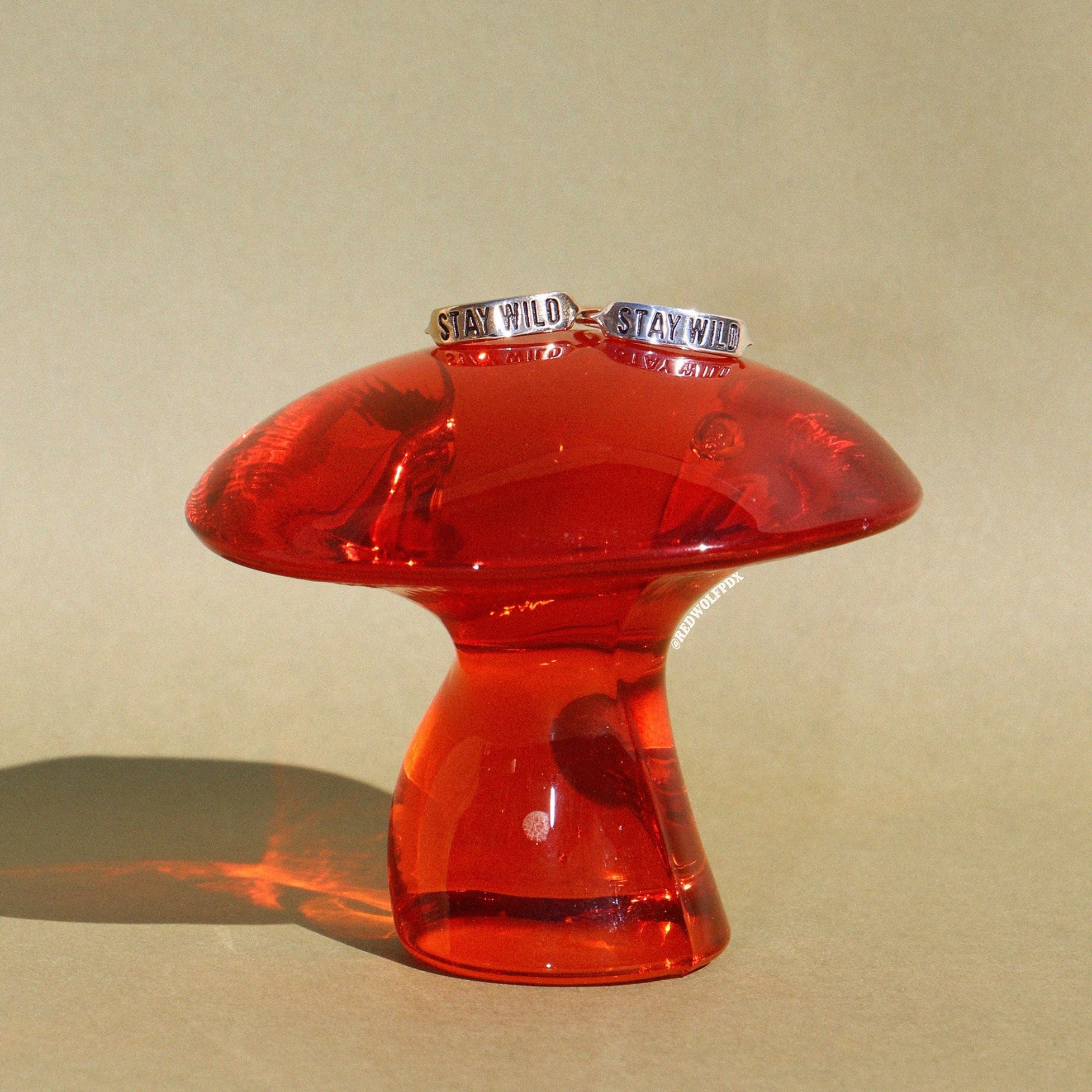 a brass ring and sterling silver ring both with text 'STAY WILD' engraved into them rested on an orange glass mushroom on a vintage green backdrop