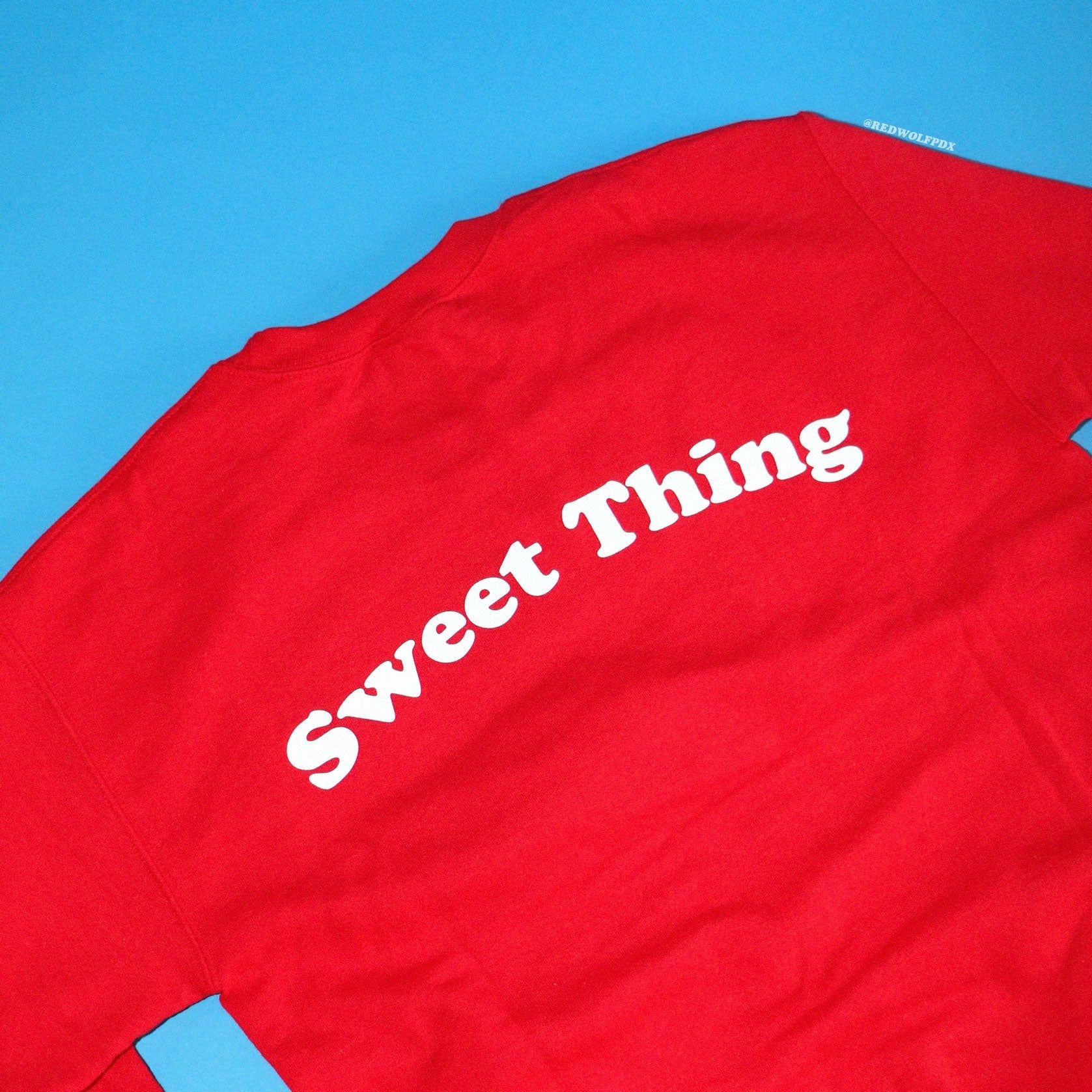 Red crewneck sweatshirt with text 'Sweet Thing' in white ink screen printed across the back against a blue background