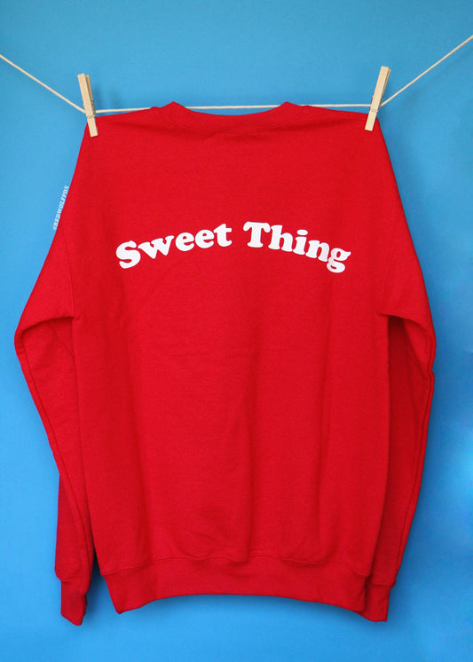 A red crewneck sweatshirt with text 'Sweet Thing' screen printed in vintage inspired font  in white ink arched text across the upper back, hanging from a clothesline with wooden clothespins against a solid blue background.