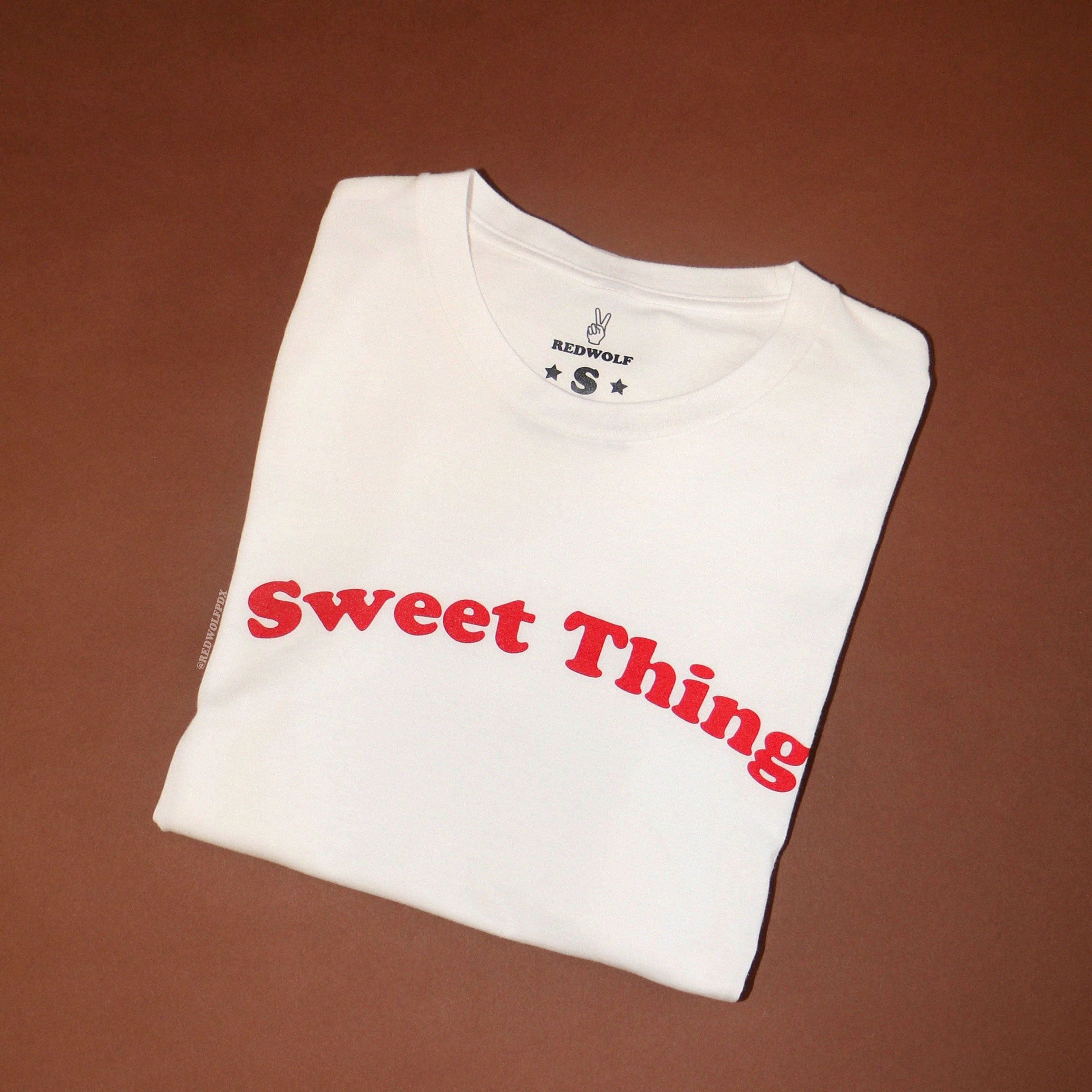 folded white graphic tee with text 'Sweet Thing' in red ink screen printed across chest on a brown background