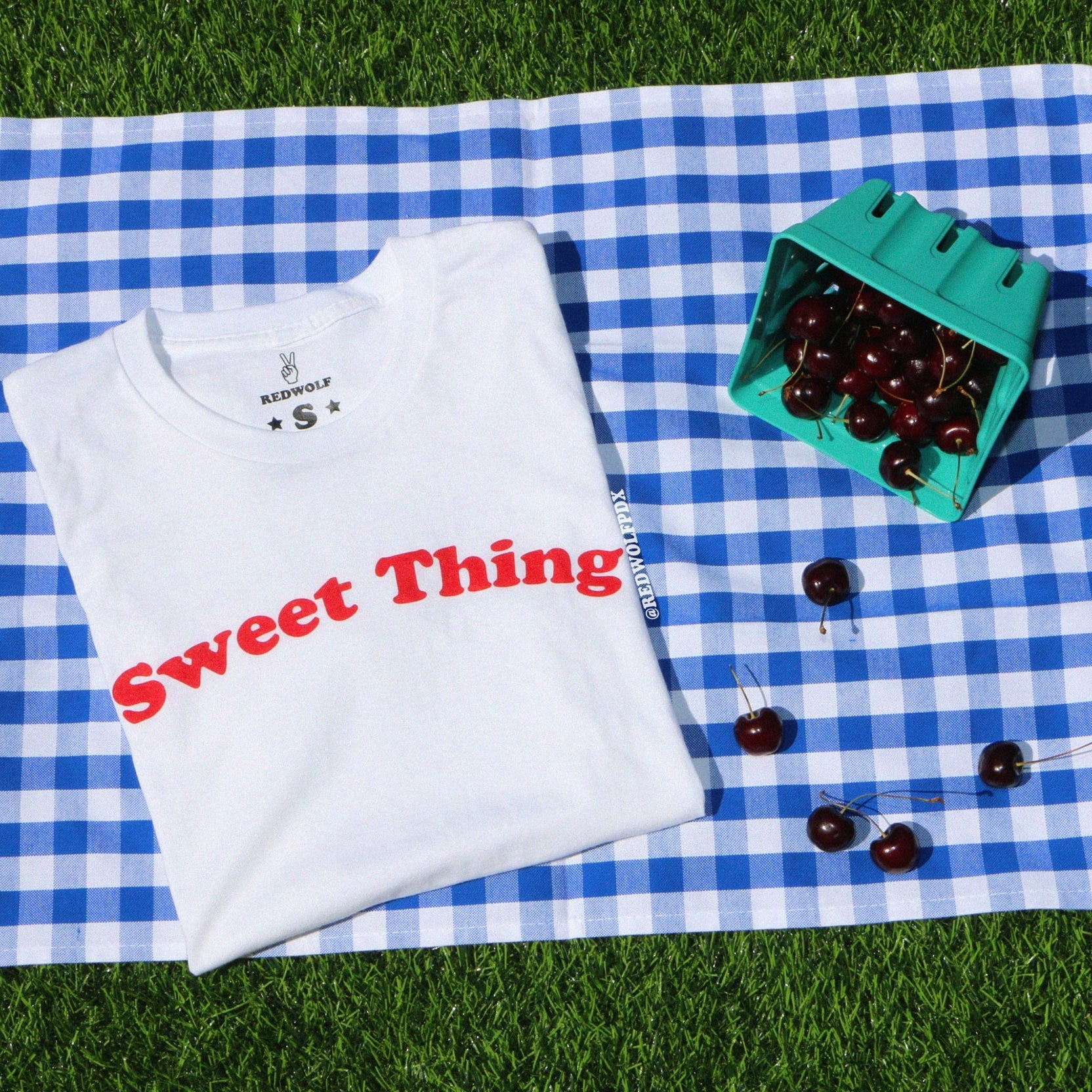 White t-shirt with 'Sweet Thing' text on a blue and white checkered blanket with  a spilled carton of cherries on green grass