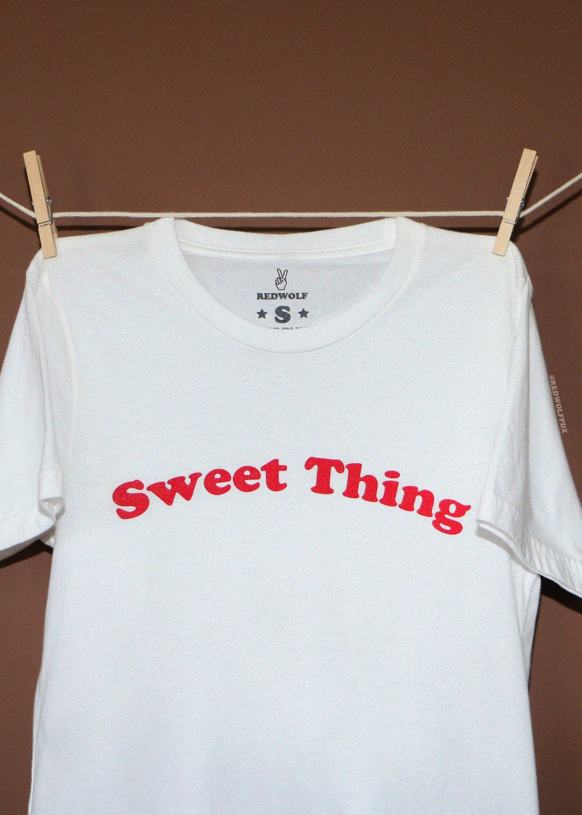 A white short-sleeve t-shirt with 'Sweet Thing' printed in red arched text across the chest, hanging from wooden clothespins on a rope against a brown background.