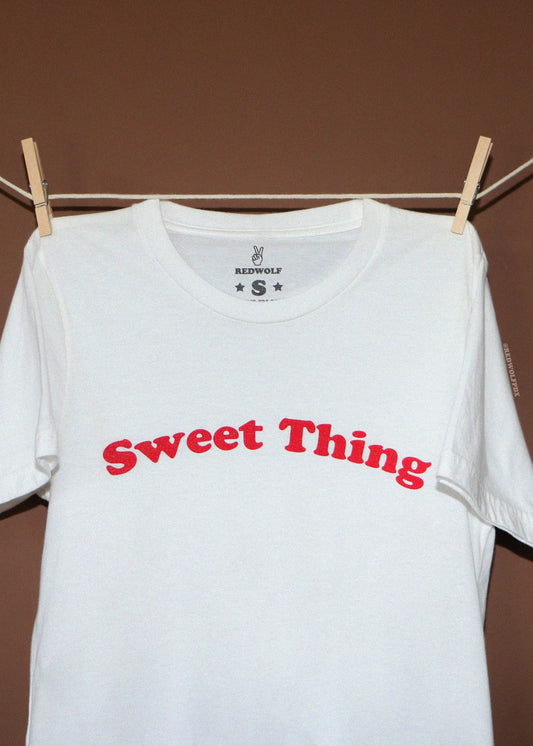A white short-sleeve t-shirt with 'Sweet Thing' printed in red arched text across the chest, hanging from wooden clothespins on a rope against a brown background.