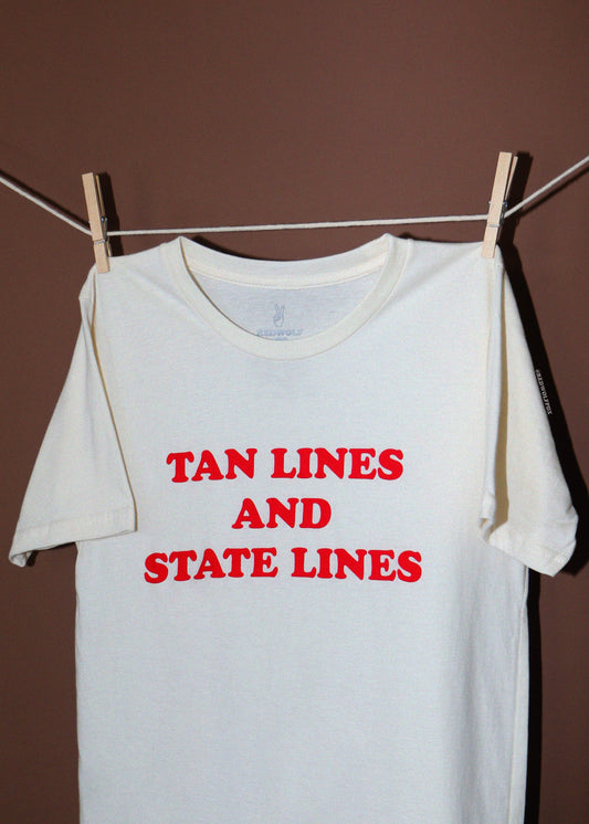 A white short-sleeve t-shirt hanging from a clothesline with wooden clothespins against a solid brown wall. The shirt features the text 'TAN LINES AND STATE LINES' printed in red capital letters across the chest.