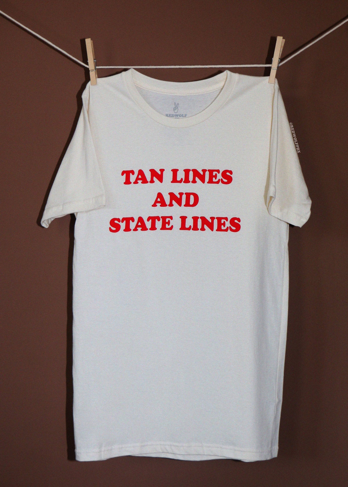 A white short-sleeve t-shirt hanging from a clothesline with wooden clothespins against a dark brown background. The shirt features the text 'TAN LINES AND STATE LINES' printed in bold red capital letters across the chest.