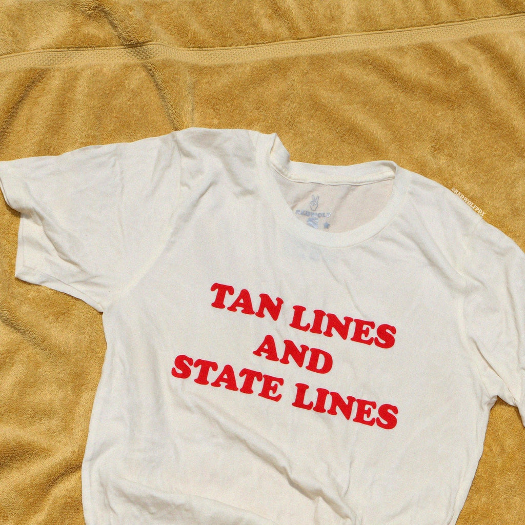 Neutral graphic tee with text 'TAN LINES AND STATE LINES' laying on golden yellow beach towel
