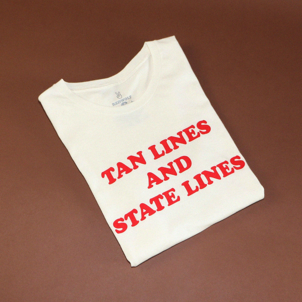 folded white tee with text 'TAN LINES AND STATE LINES' in red ink screen printed across chest on brown background.