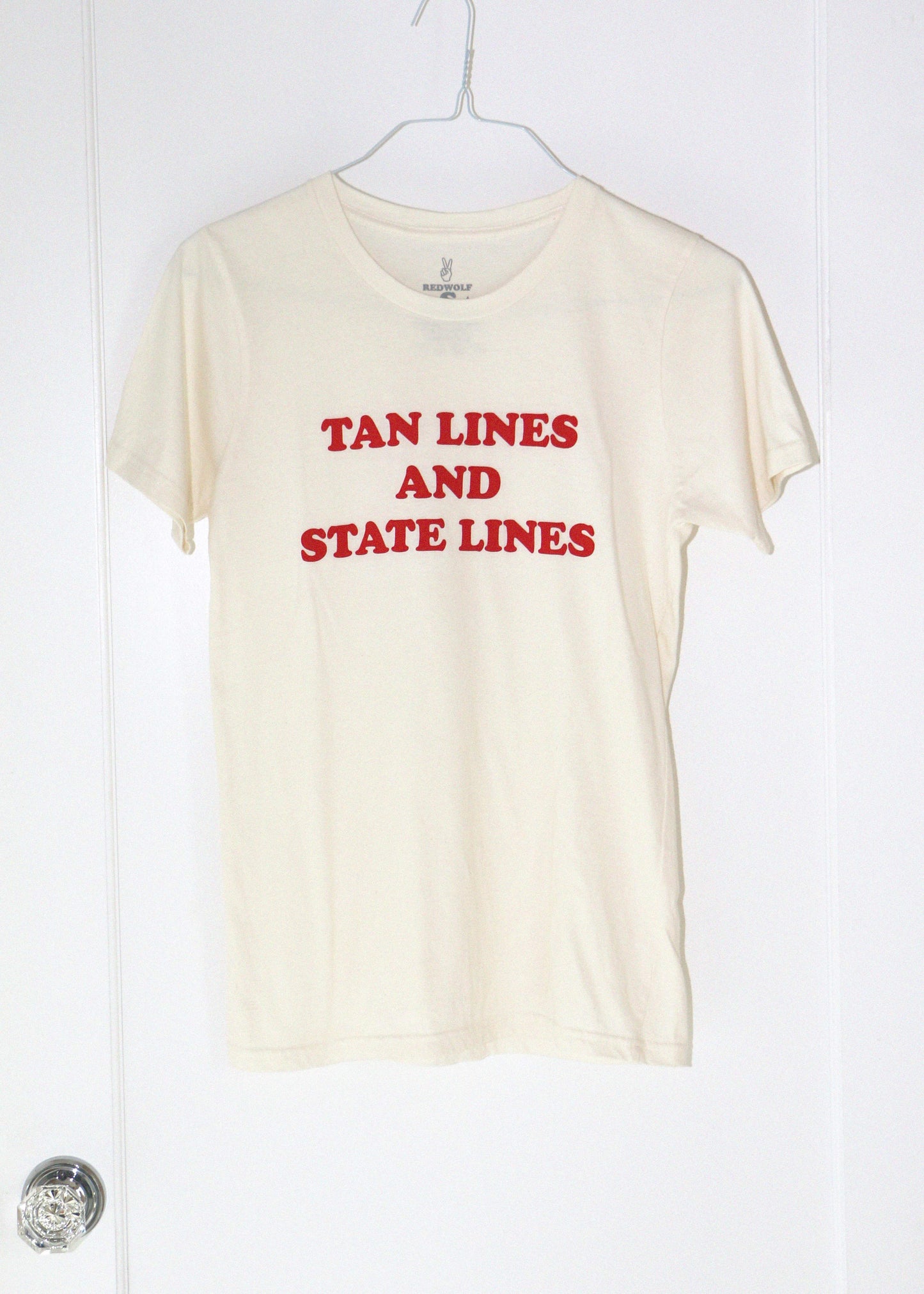 Beige t-shirt with red text 'TAN LINES AND STATE LINES' on a white hanger against a white background.