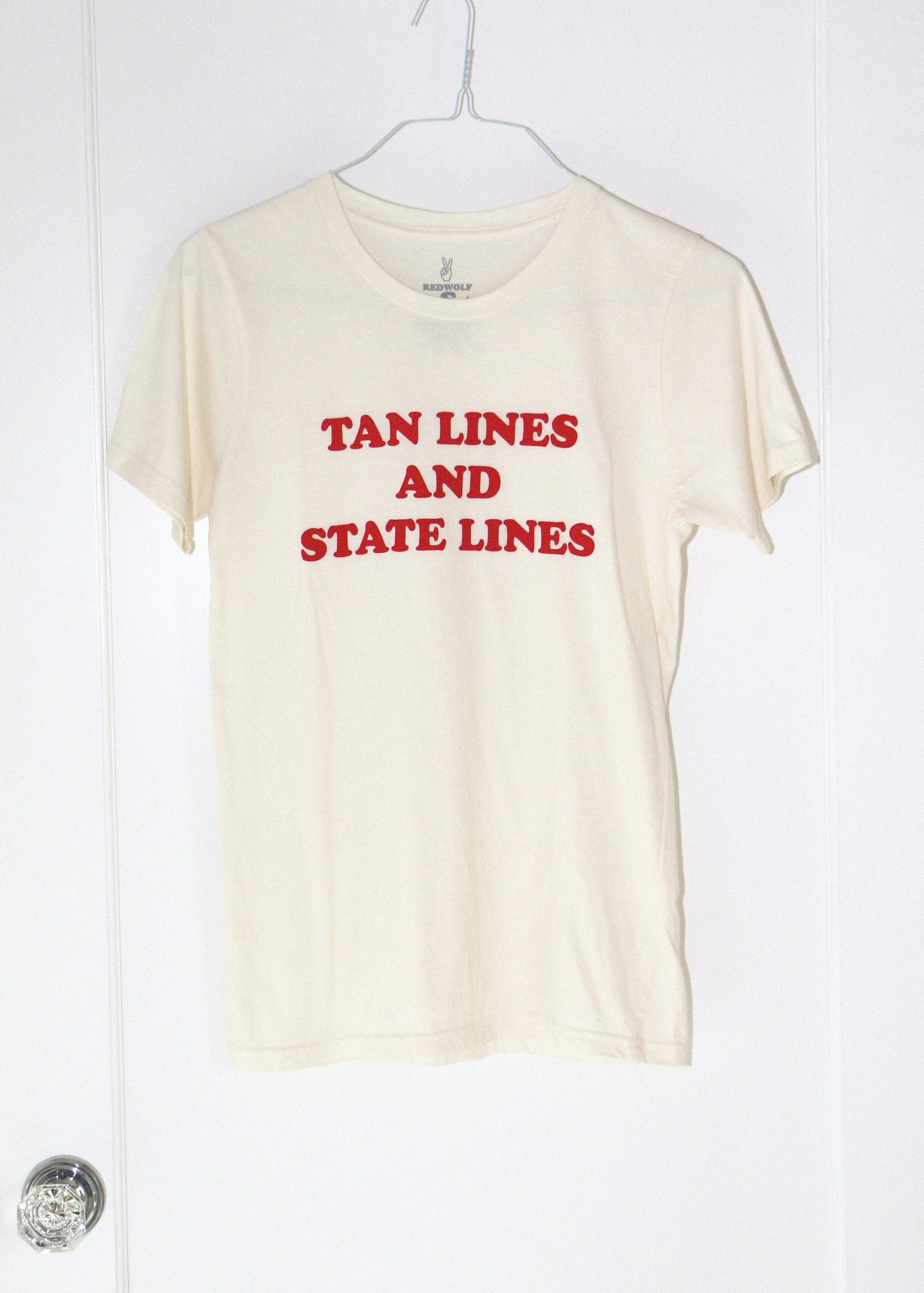 Beige t-shirt with red text 'TAN LINES AND STATE LINES' on a white hanger against a white background.