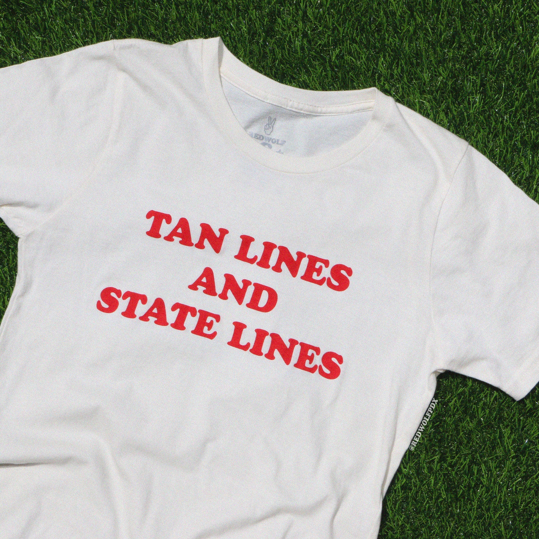 white graphic tee with text 'TAN LINES AND STATE LINES' in red ink screen printed across chest  against green grass backdrop