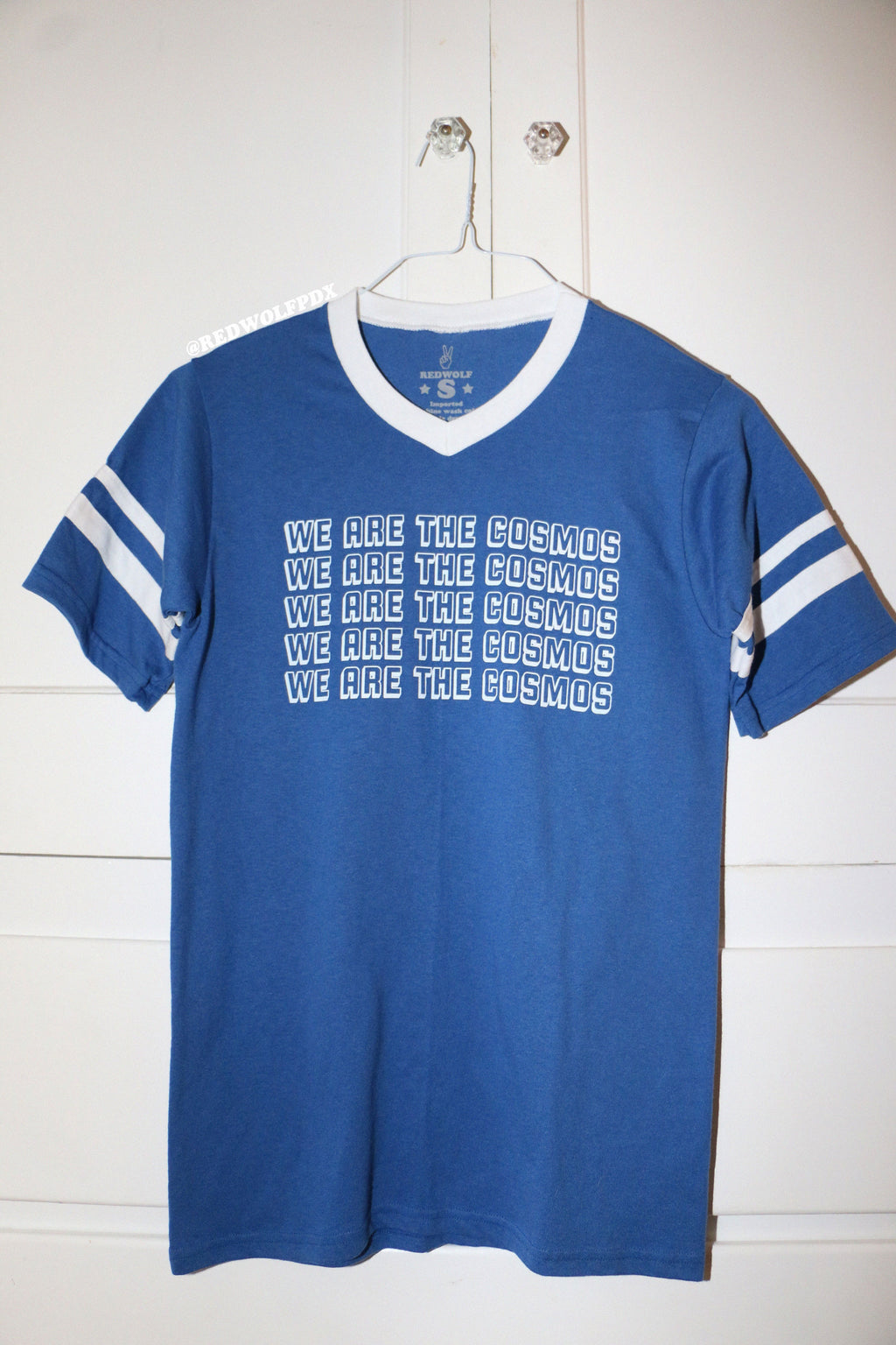Blue t-shirt with 'We Are The Cosmos' text hanging from a hanger on closet door knob