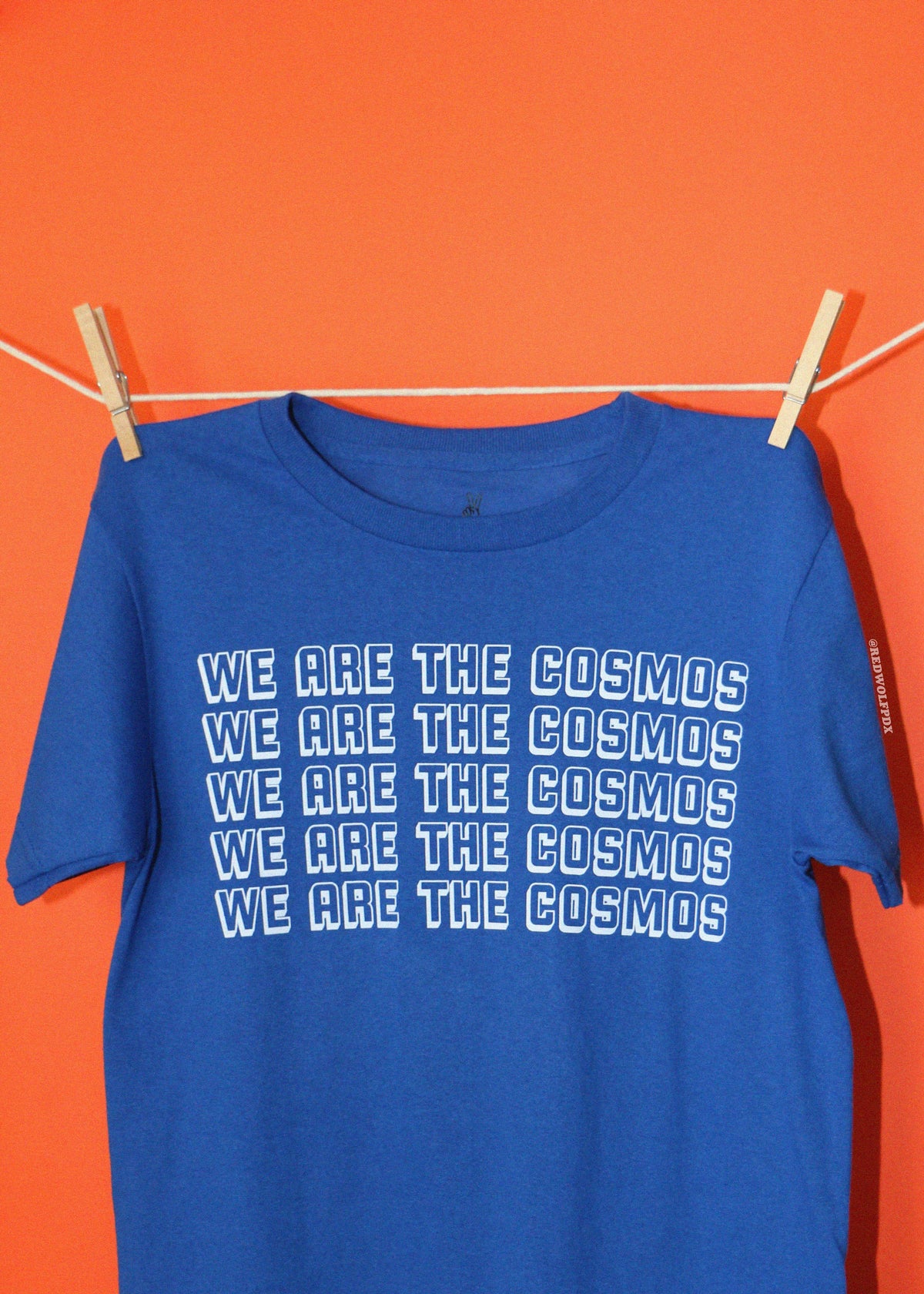 A blue short-sleeve t-shirt with the phrase 'WE ARE THE COSMOS' printed in white outlined text, hanging from a clothesline with wooden clothespins against a solid orange background.
