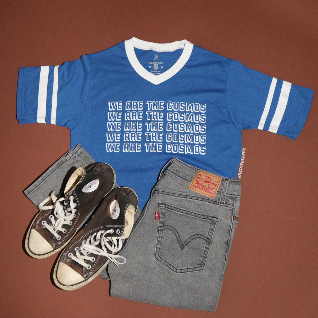 Blue t-shirt with 'We Are The Cosmos' text  with black jeans and sneakers flatlay on brown background