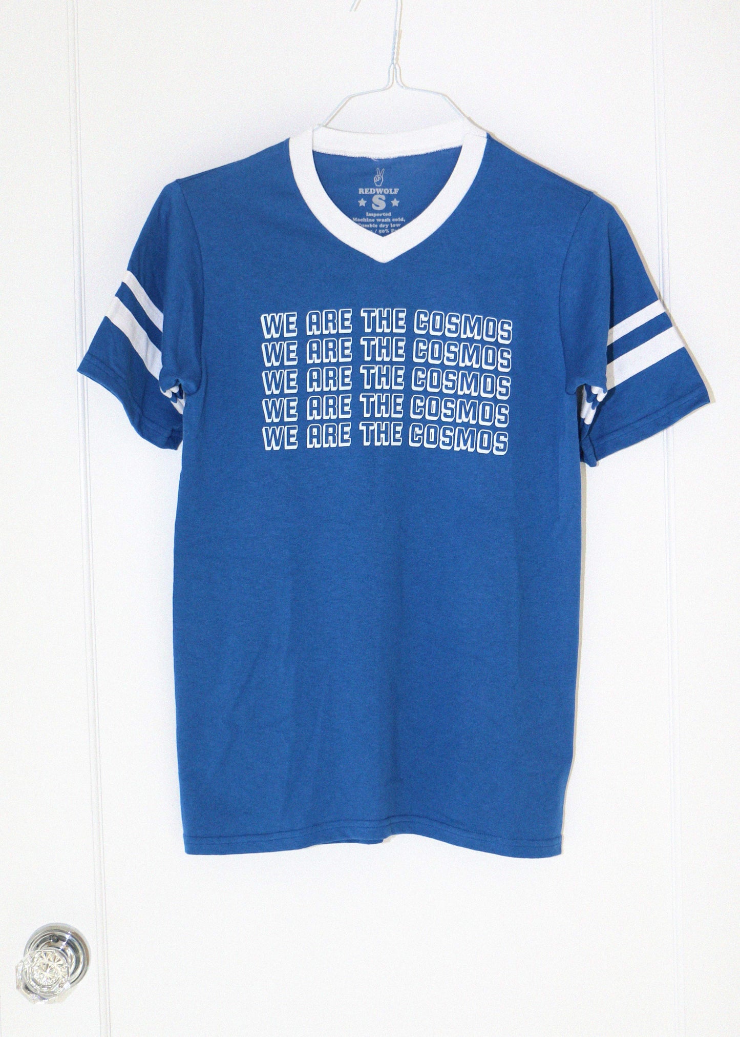 Blue t-shirt with white text 'we are the cosmos' and stripes on a hanger against a white background