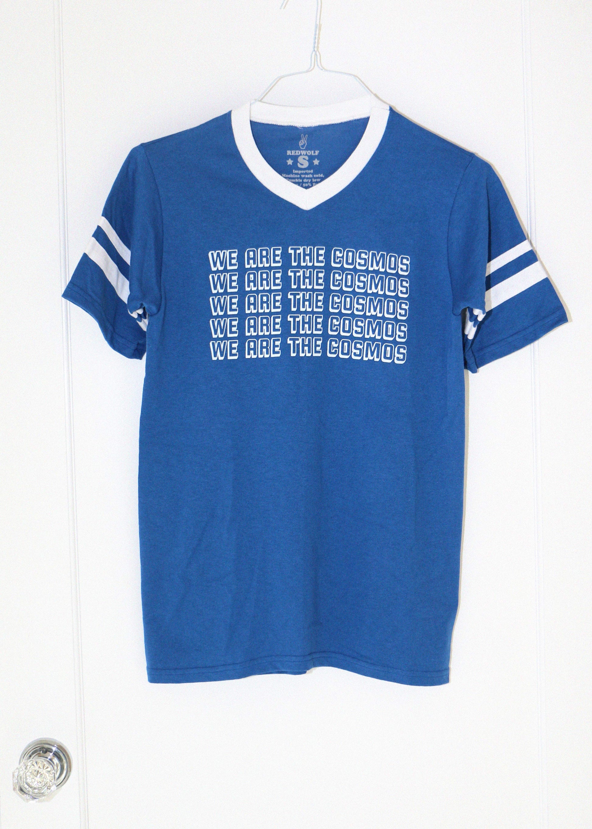 Blue t-shirt with white text 'we are the cosmos' and stripes on a hanger against a white background