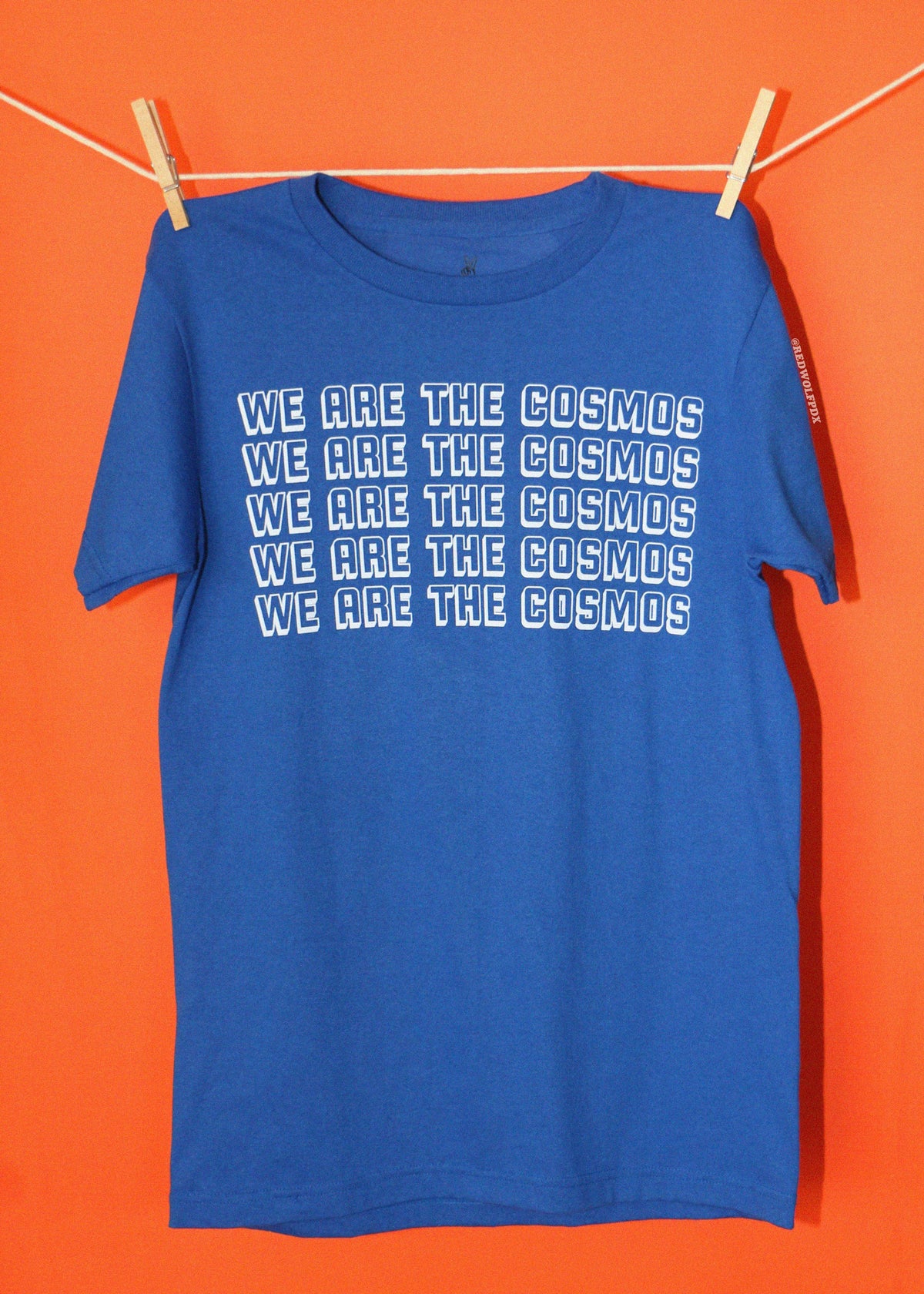 A blue short-sleeve t-shirt hanging from a clothesline with wooden clips against a solid orange background. The shirt features the phrase 'WE ARE THE COSMOS' repeated in white outline text across the chest.