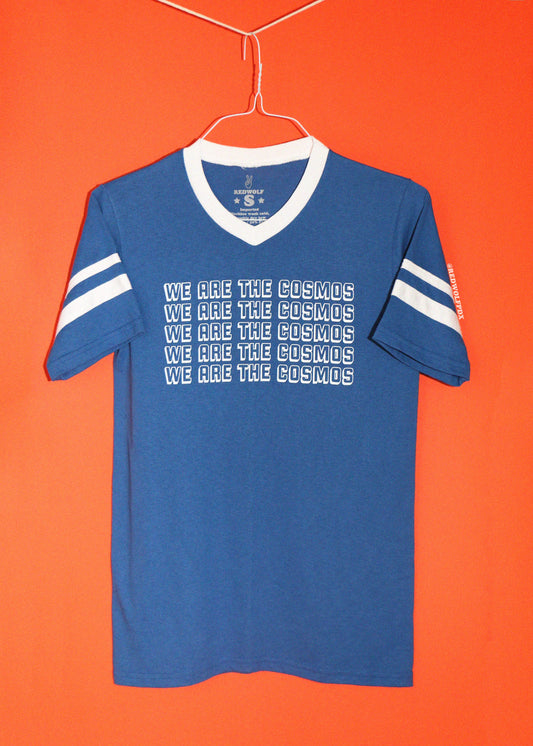 A retro inspired blue v-neck t-shirt with white stripes on the sleeves and white text reading 'WE ARE THE COSMOS' repeated across the front, hanging on a wire hanger against a solid orange background.