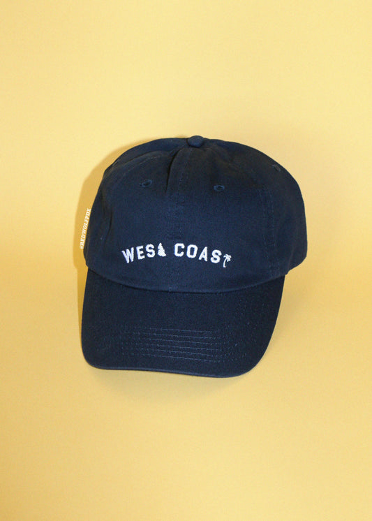 A navy blue baseball cap with 'WEST COAST' embroidered in white on the front, displayed against a solid pale yellow background.