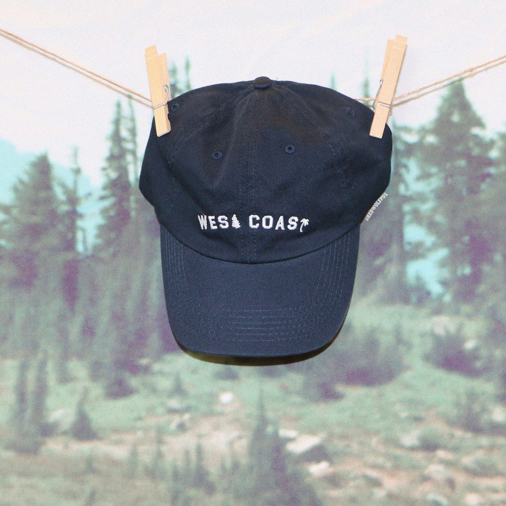 Navy blue baseball cap with text 'WEST COAST' embroidered with white thread hanging from wood clothings pins with a nature forest backdrop