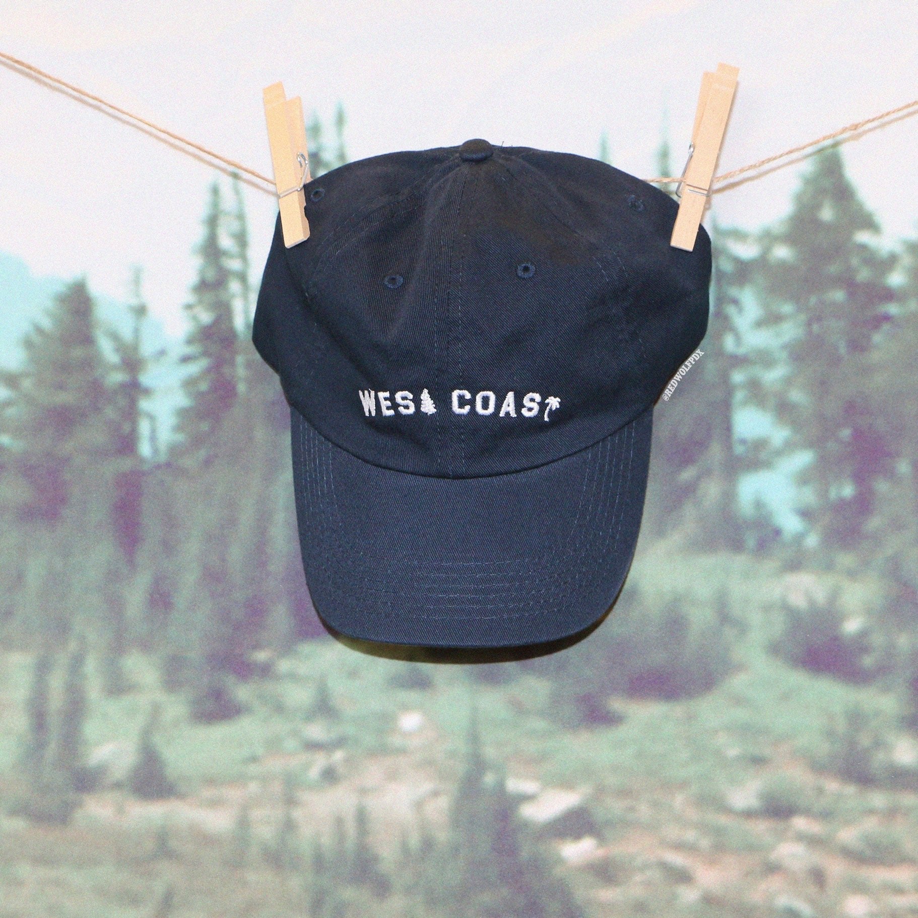 Navy blue baseball cap with text 'WEST COAST' embroidered with white thread hanging from wood clothings pins with a nature forest backdrop