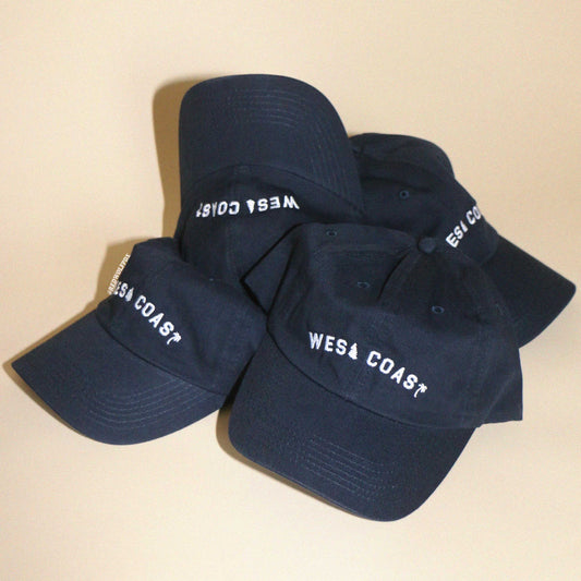Pile of navy blue baseball caps with text 'WEST COAST' embroidered in white thread on beige backdrop. 