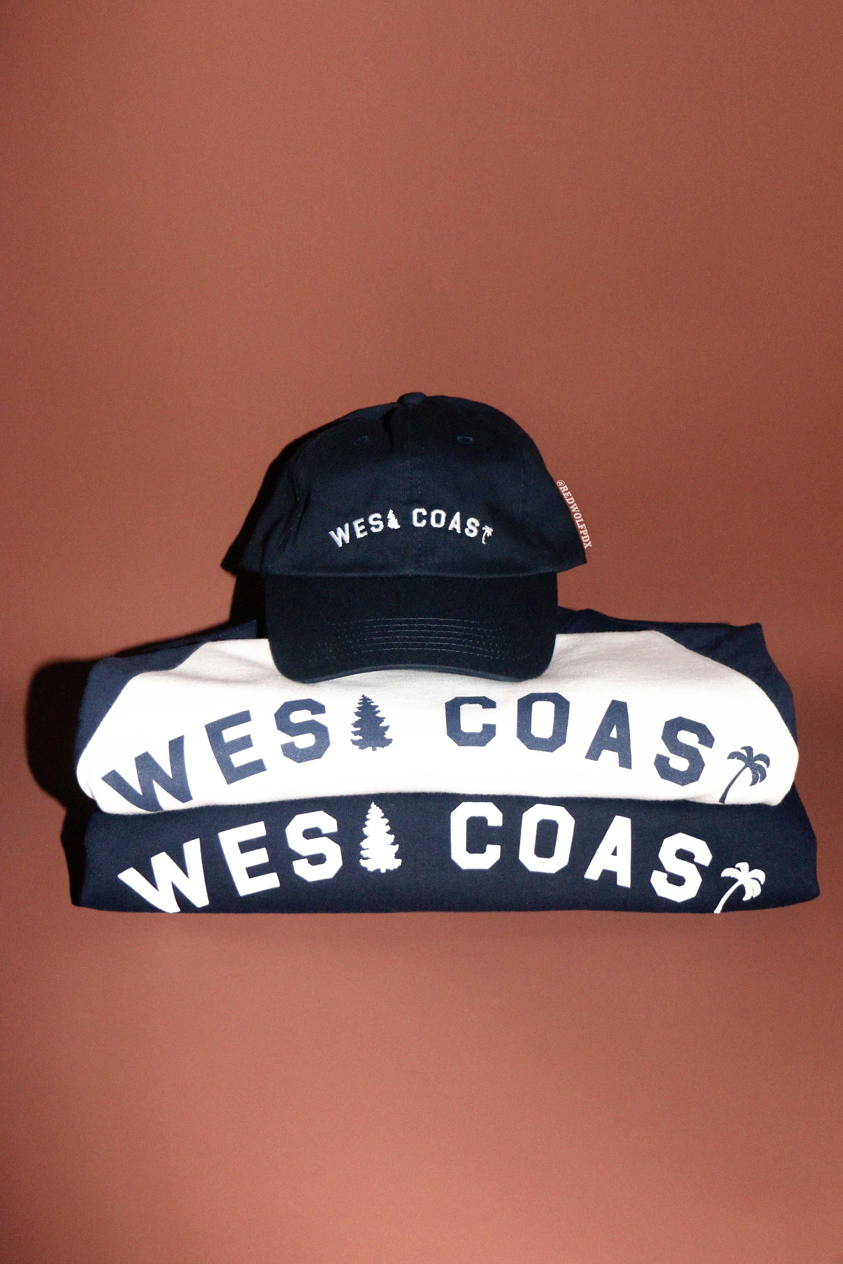 navy blue baseball cap stacked on white tee and navy blue crewneck sweatshirt all with 'WEST COAST' text on a brown background