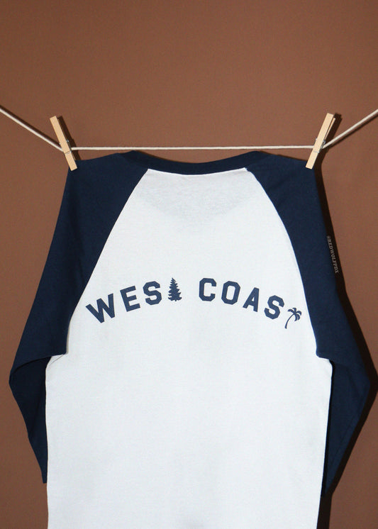 A raglan-style baseball shirt hanging from a clothesline with wooden clothespins against a solid brown background. The shirt has a white body and navy blue sleeves, with 'WEST COAST' printed in navy blue across the back.