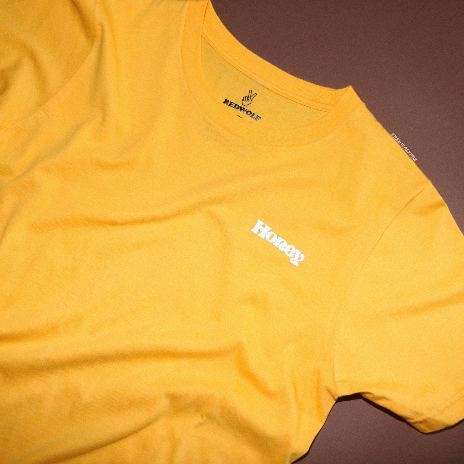 bright mustard yellow graphic tee with text 'Honey' in white ink screen printed on corner of chest on a brown background