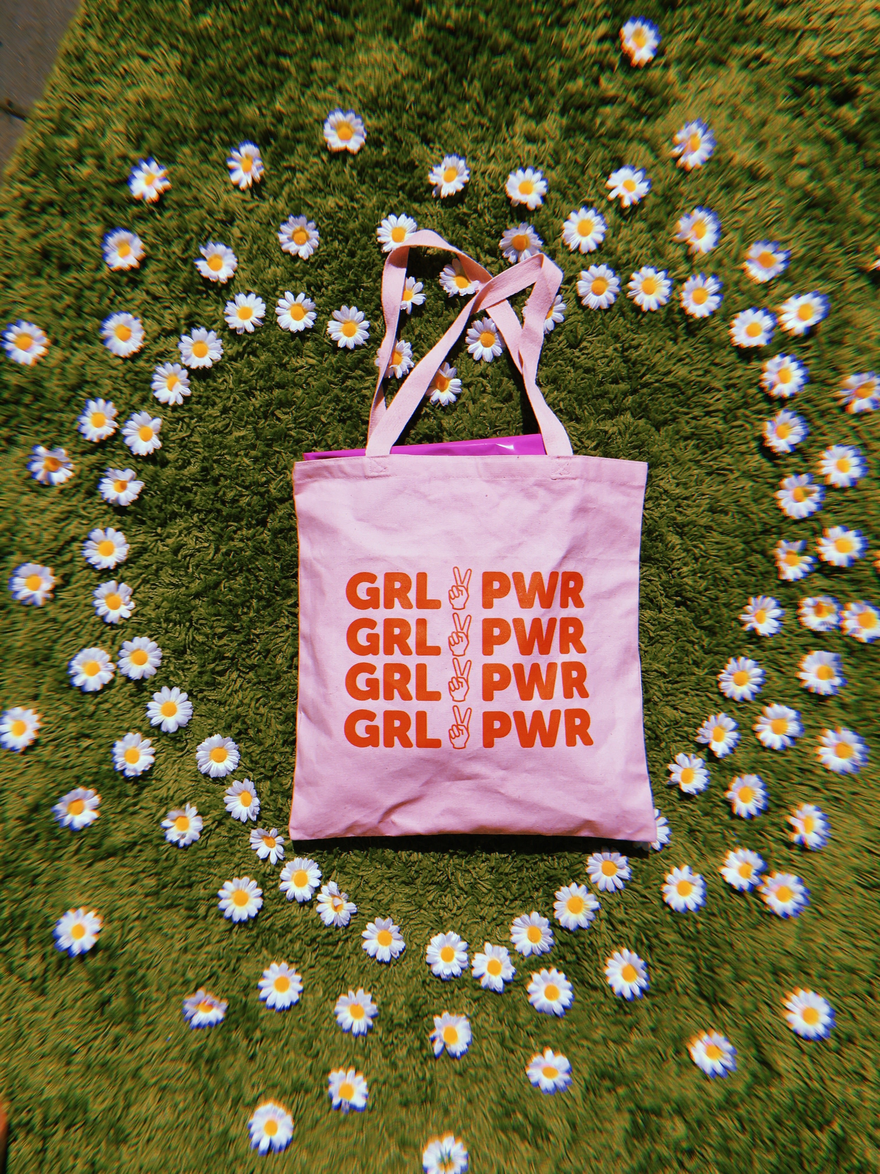 light pink canvas tote bag with text 'GRL PWR' in red orange ink screen printed down front against a green shaggy rug and surrounded by daises in shape of a heart