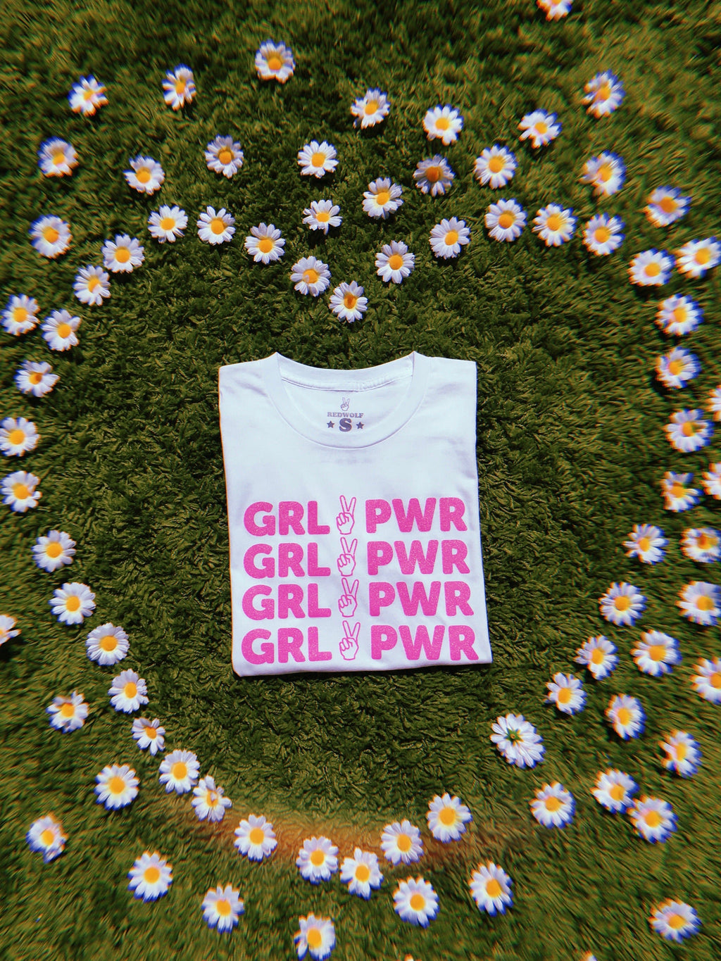 folded white tee with text 'GRL PWR' with peace sign graphic screen printed in pink glitter ink across the front on  a green shaggy rug with daisies scattered in shape of heart framing the tee