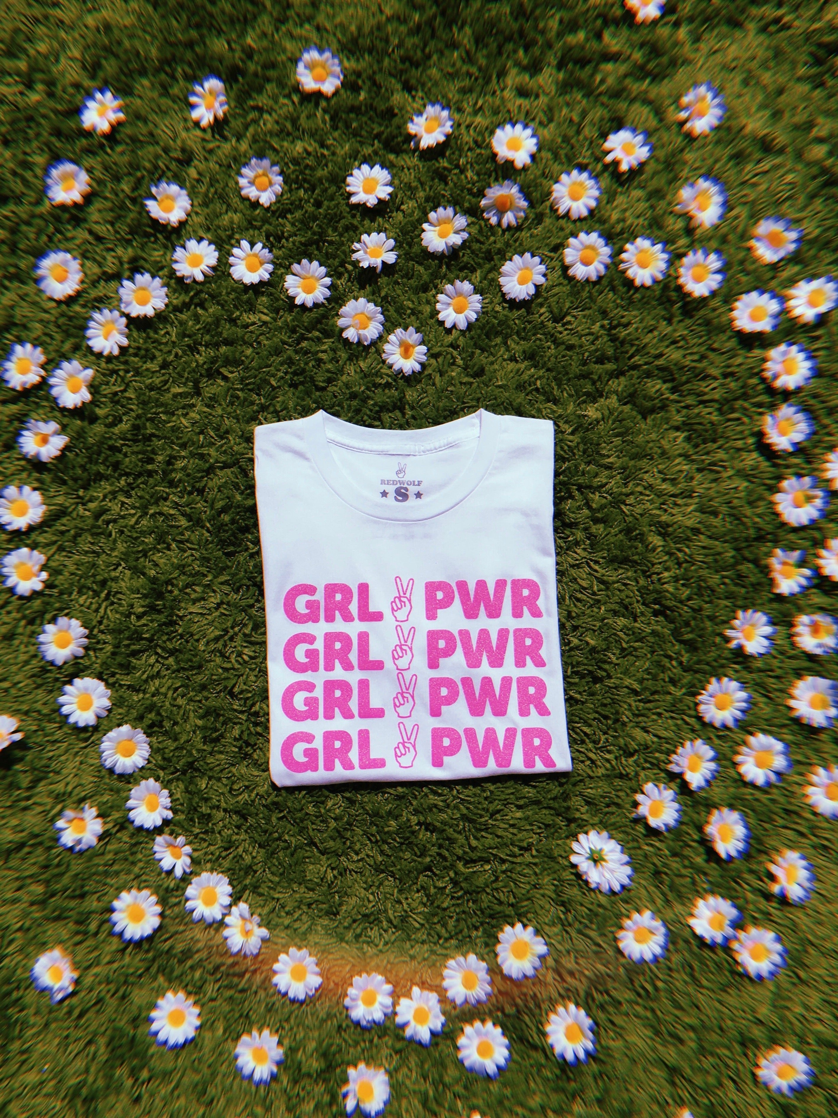 folded white tee with text 'GRL PWR' with peace sign graphic screen printed in pink glitter ink across the front on  a green shaggy rug with daisies scattered in shape of heart framing the tee