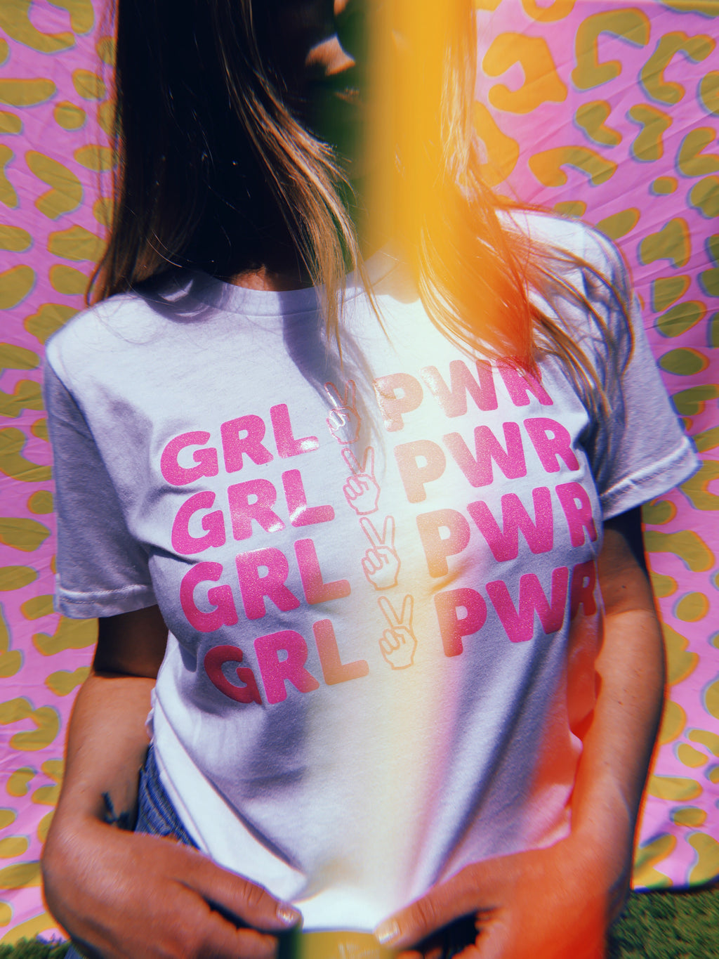 white tee with text 'GRL PWR' with peace sign graphic screen printed in pink glitter ink across the front being worn against a colorful background