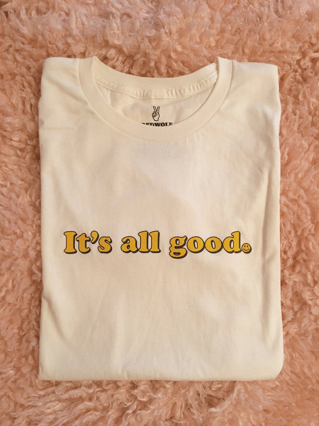 beige folded graphic tee with text 'It's all good' with smiley face illustration in yellow ink screen printed across chest on a pink shaggy rug background