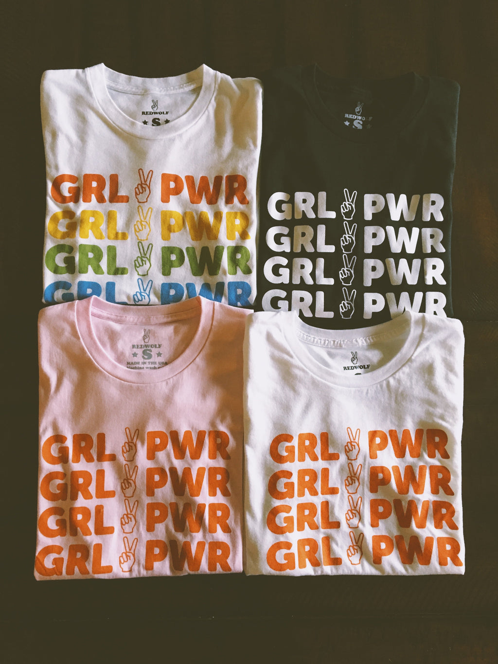 folded tees of different colors with text 'GRL PWR' and peace sign graphic screen printed down the front on a wood background