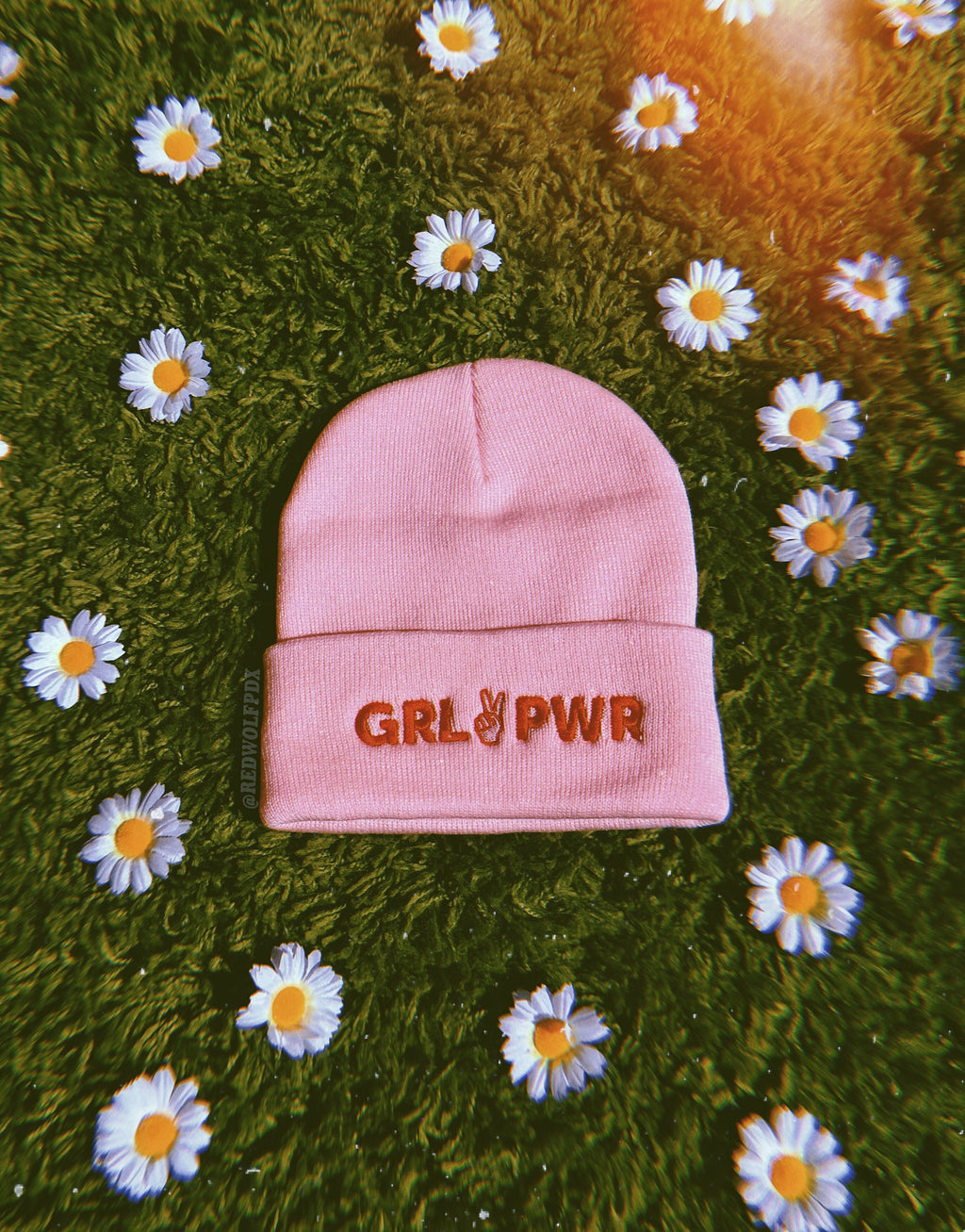 light pink beanie with text 'GRL PWR' in red thread embroidered across band of the beanie against green shaggy rug and daisies