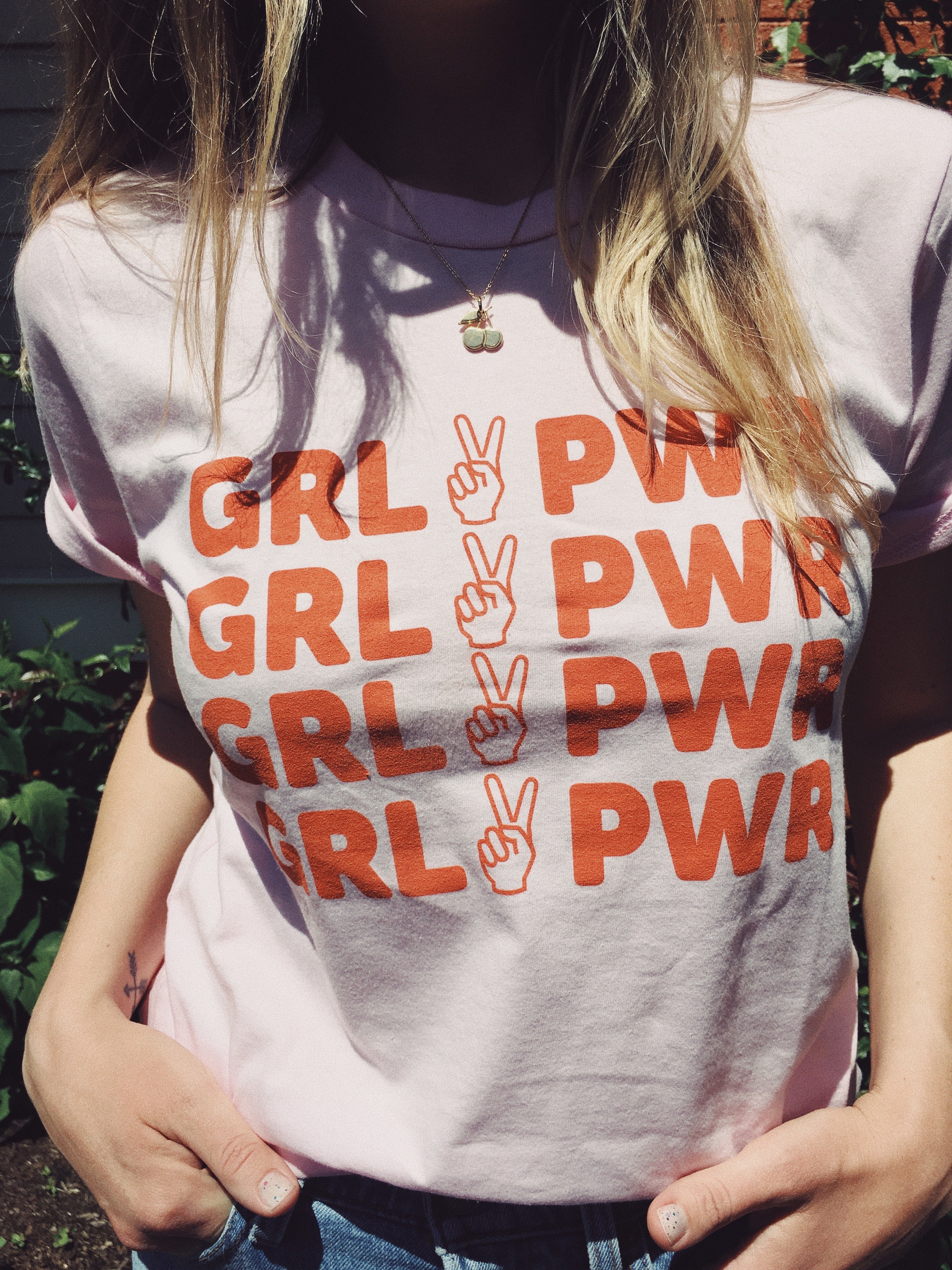 pink tee with text 'GRL PWR' with peace sign graphic in orange/red ink screen printed down front being worn in yard