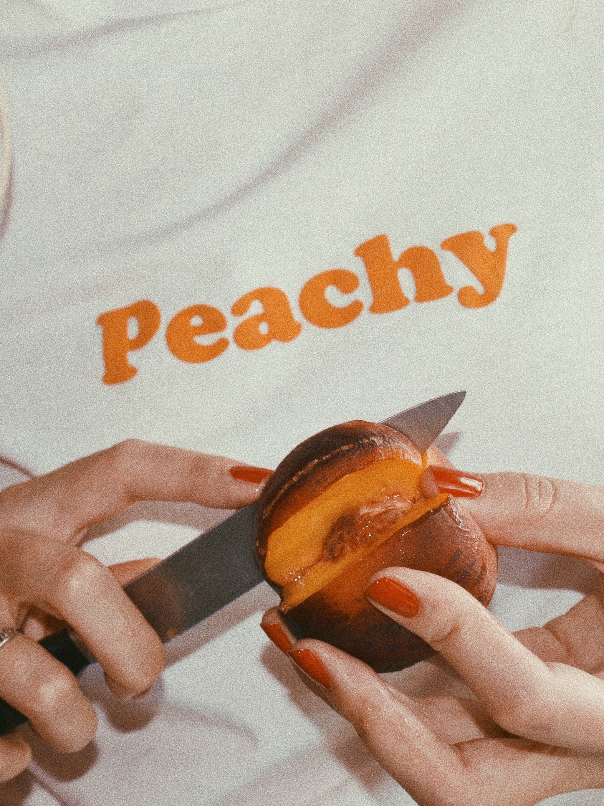 white graphic tee with text 'Peachy' in orange ink screen printed on chest being worn while being worn slicing a peach with a knife
