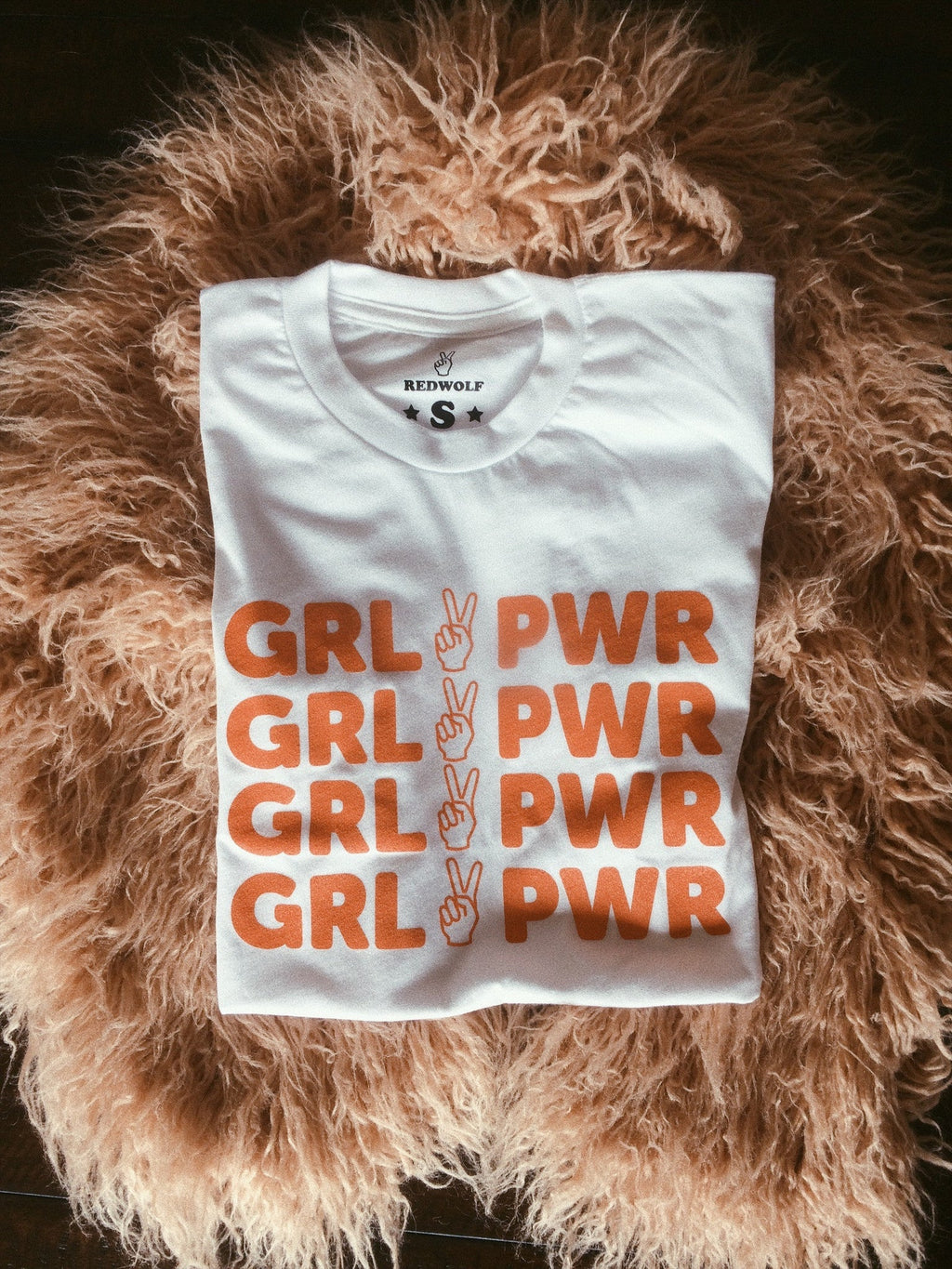 Folded white tee with text 'GRL PWR' with peace sign graphic in vintage red orange ink screen printed down front on a pink faux fur background